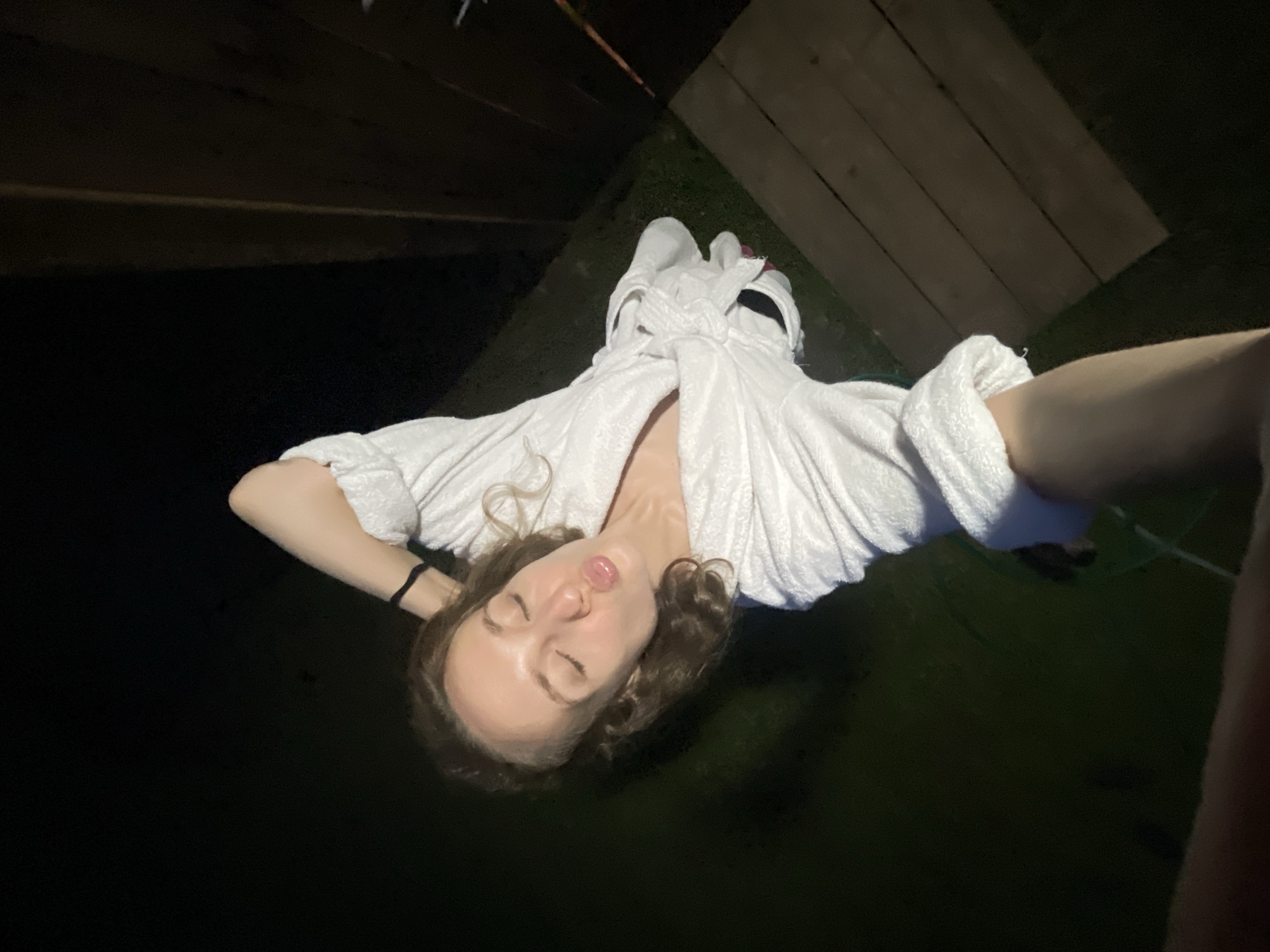 The media file shows a person lying down outdoors at night, possibly in Sharkan, Russia. The individual is wearing a white bathrobe and has their eyes closed, with their lips slightly pursed as if making a kissing gesture. Their arms are behind their head, and their hair is a light brown color. The scene is dark, illuminated by artificial light, suggesting it is nighttime. In the background, there are wooden planks and some green ground, possibly grass, indicating an outdoor setting like a yard or patio. The overall mood appears relaxed and playful. There is no visible text in the image.