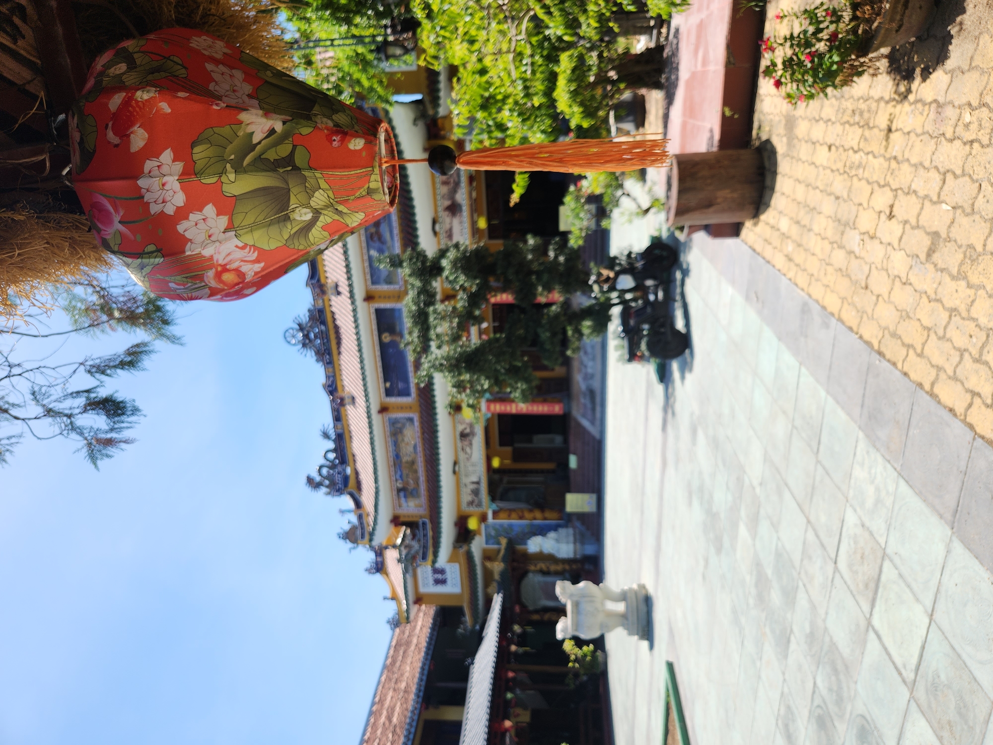 A red lantern with lotus and koi fish motifs hangs from a thatched roof. In the background, a yellow temple with a tiled roof and intricate decorations is visible. The temple grounds are paved with light-colored tiles and a yellow patterned border. A white ornamental urn is positioned on the left side of the courtyard, and a three-wheeled vehicle is parked in the middle ground. Lush green trees and foliage surround the temple and courtyard. The scene is set on a sunny day with a clear blue sky. The location is a temple in Hoi An, Vietnam.