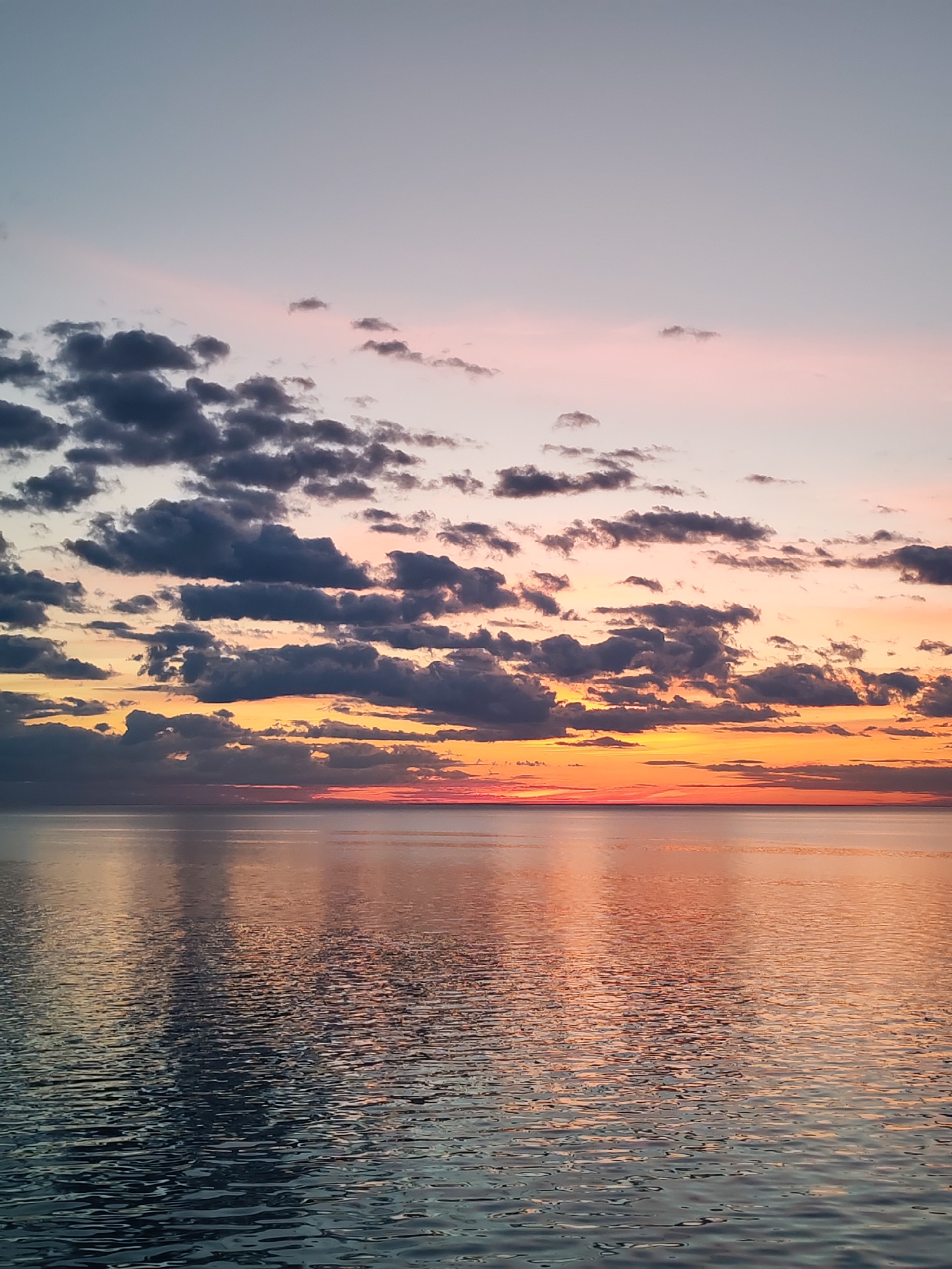 The image depicts a seascape at sunset in Bol'shie Mkhi, Russia. The sky transitions from a pale blue and pink at the top to a vibrant orange and red near the horizon. Numerous dark gray clouds are scattered across the sky. Below the sky is a body of water, likely a lake or sea, reflecting the colors of the sunset. The water has small ripples and waves, indicating a gentle breeze. The overall scene is calm and serene, with a focus on the natural beauty of the sunset and its reflection on the water.