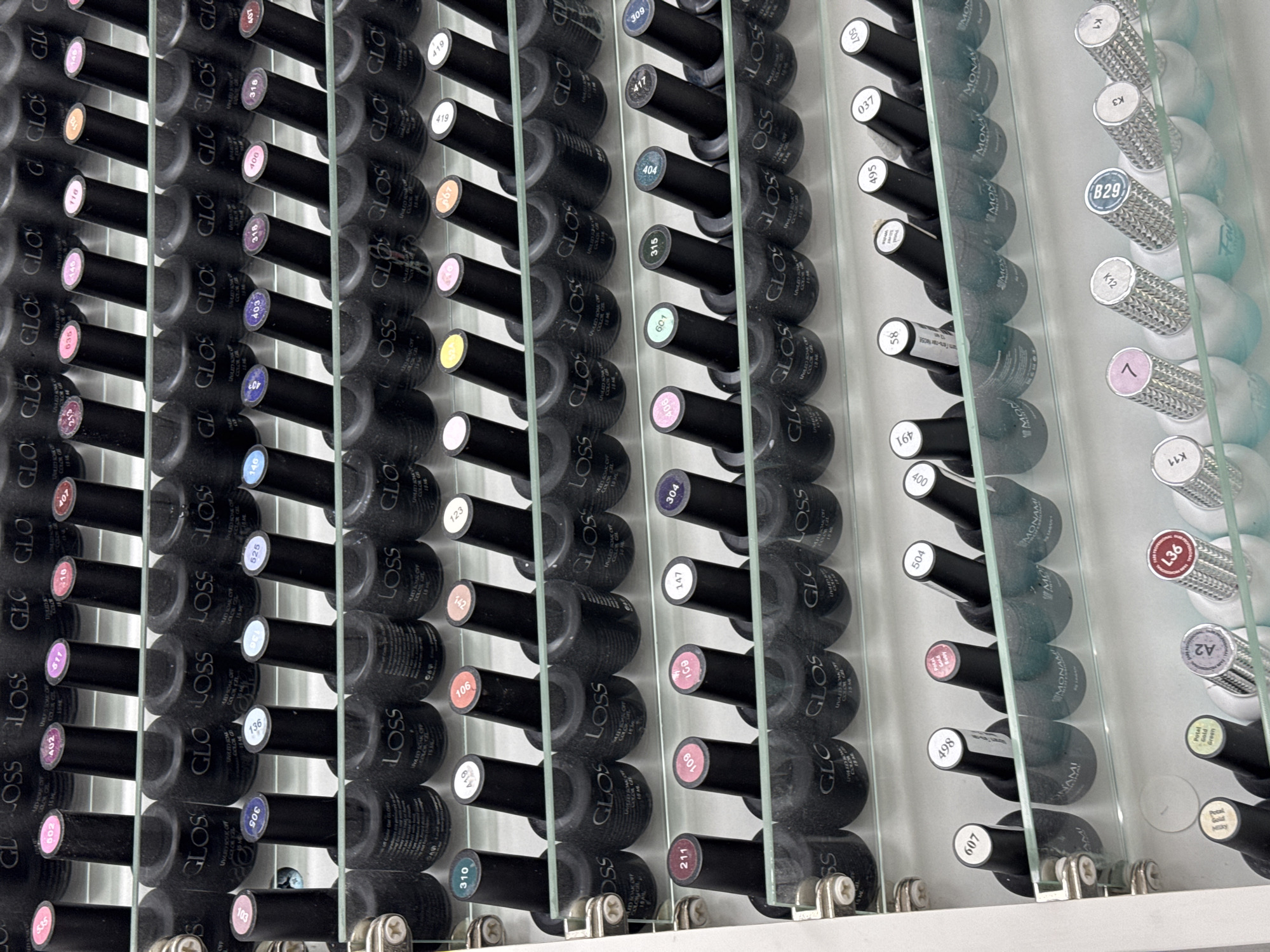 The image displays a wall of nail polish bottles organized in rows and columns. The bottles are primarily black with colorful caps indicating the shade of polish. Many bottles have labels with numbers, and some also show brand names like "GLOSS," "MONAMI," and "COLOR." The bottles are stored on clear shelves, with a reflective surface behind them, suggesting a retail or salon environment. Some of the labels are clearly legible, showing numbers such as "511," "402," "602," "400," "419," "106," "123," "304," "147," "211," "109," "601," "315," "404," "406," "498," "504," "607," "58," "495," "037," "209," "491." Other labels include "GLOSS," "COLOR GEL," "UNALTY SOAK OFF," "UVLED SOAK OFF," "MONAMI," "B29," "K12," "K3," "K11," "A2," and "7." The overall impression is one of an organized and well-stocked display of nail care products. The setting appears to be indoors, possibly a beauty salon or a retail store.