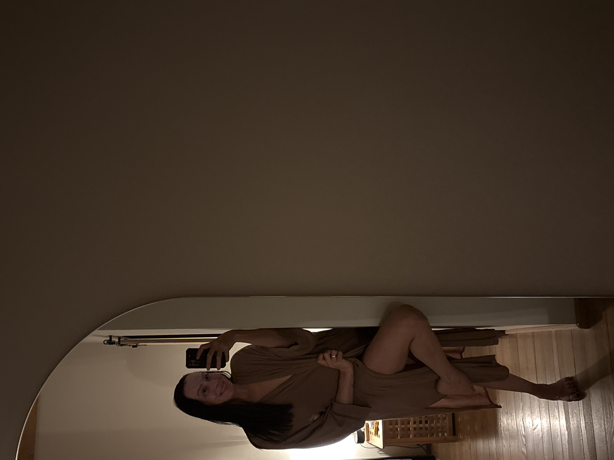 A woman is taking a selfie in a mirror. She is wearing a brown robe and is lying down. The mirror is hanging on a wall and reflects the room. There is a nightstand to the right of the woman. The floor is made of wooden planks. It is not possible to discern the location as Chaikovsky, Russia from the image.