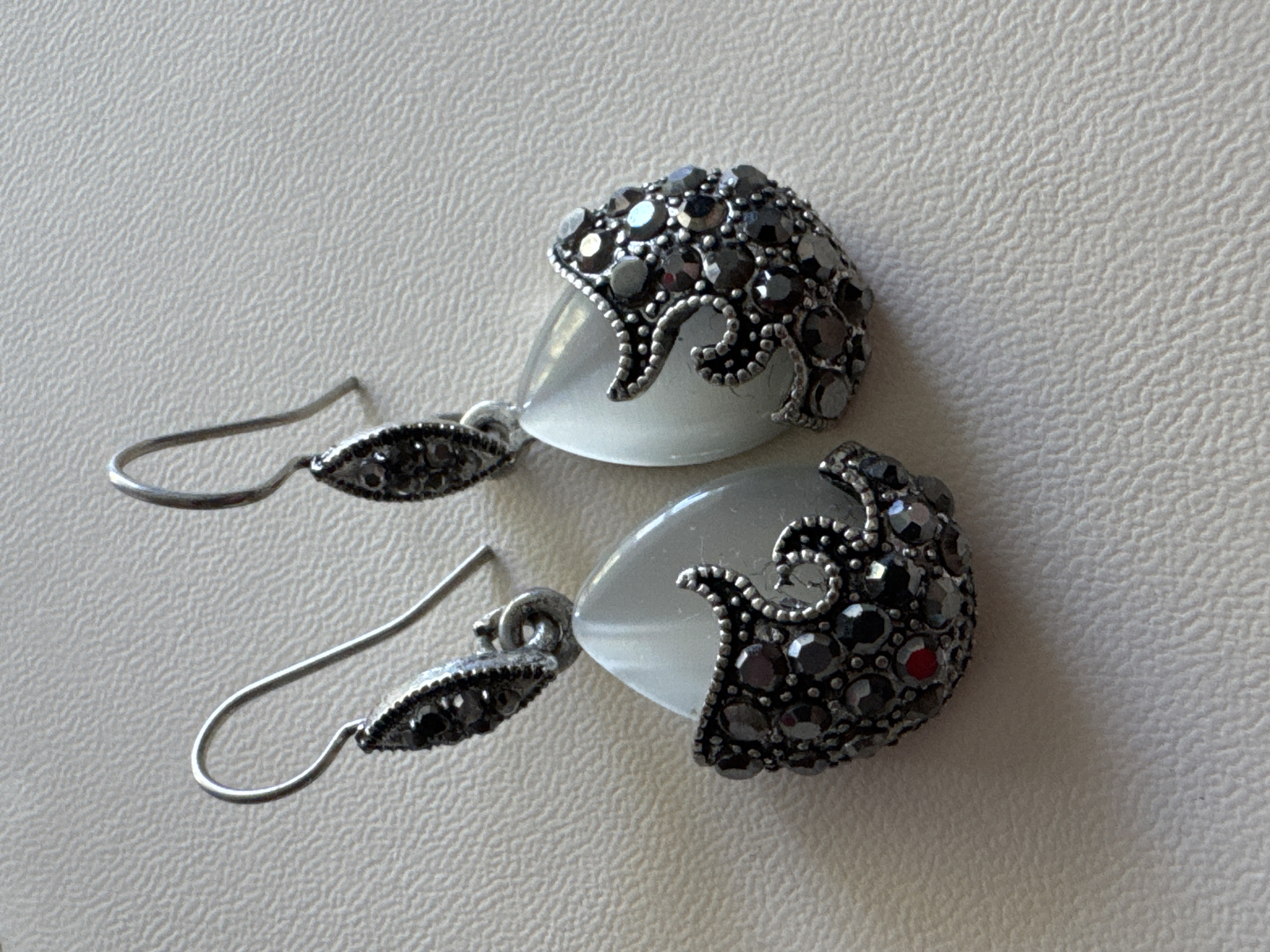 A pair of ornate silver earrings, each featuring a teardrop-shaped milky white stone surrounded by dark, faceted stones and intricate silver detailing, are displayed on a textured, light-colored surface. The earrings are positioned close together, with the hooks of each earring visible and pointing upwards. The surface they are on appears to be a placemat or fabric with a subtle, repeating pattern. The lighting suggests an indoor environment, possibly a studio or home setting. There are no people or any other objects visible in the scene. The earrings are the sole focus of the image. The background is a neutral, textured surface.