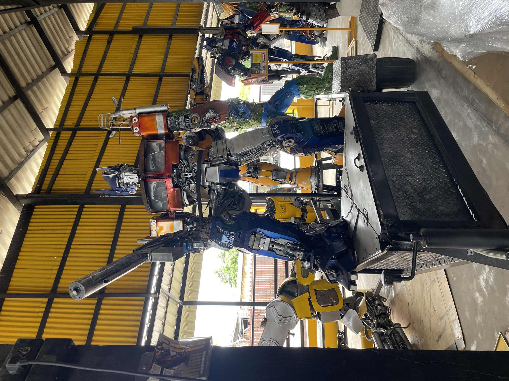An elaborate sculpture of the Transformer character Optimus Prime, constructed from scrap metal, stands prominently in a workshop or exhibition space. The sculpture is detailed, featuring its signature red and blue coloration, and is posed dynamically with a large weapon. To the left and right of Optimus Prime, other Transformer-like sculptures, also made from repurposed materials, are visible, including what appears to be Bumblebee and another character with green legs and blue torso. The background is characterized by a corrugated yellow metal wall and a metal roof structure, typical of industrial or open-air buildings. The flooring is concrete, with various pieces of metal and other materials scattered around, suggesting an active workshop environment. In the foreground, a metal trailer, also constructed from scrap metal, is positioned in front of Optimus Prime, with a diamond plate pattern on its sides and a wheel. The overall impression is one of creative reuse of materials to form impressive and large-scale robotic figures. The location is identified as Pattaya City, Thailand, hinting at a possible tourist attraction or a local artisan's workshop. There are no people visibly present in the image.