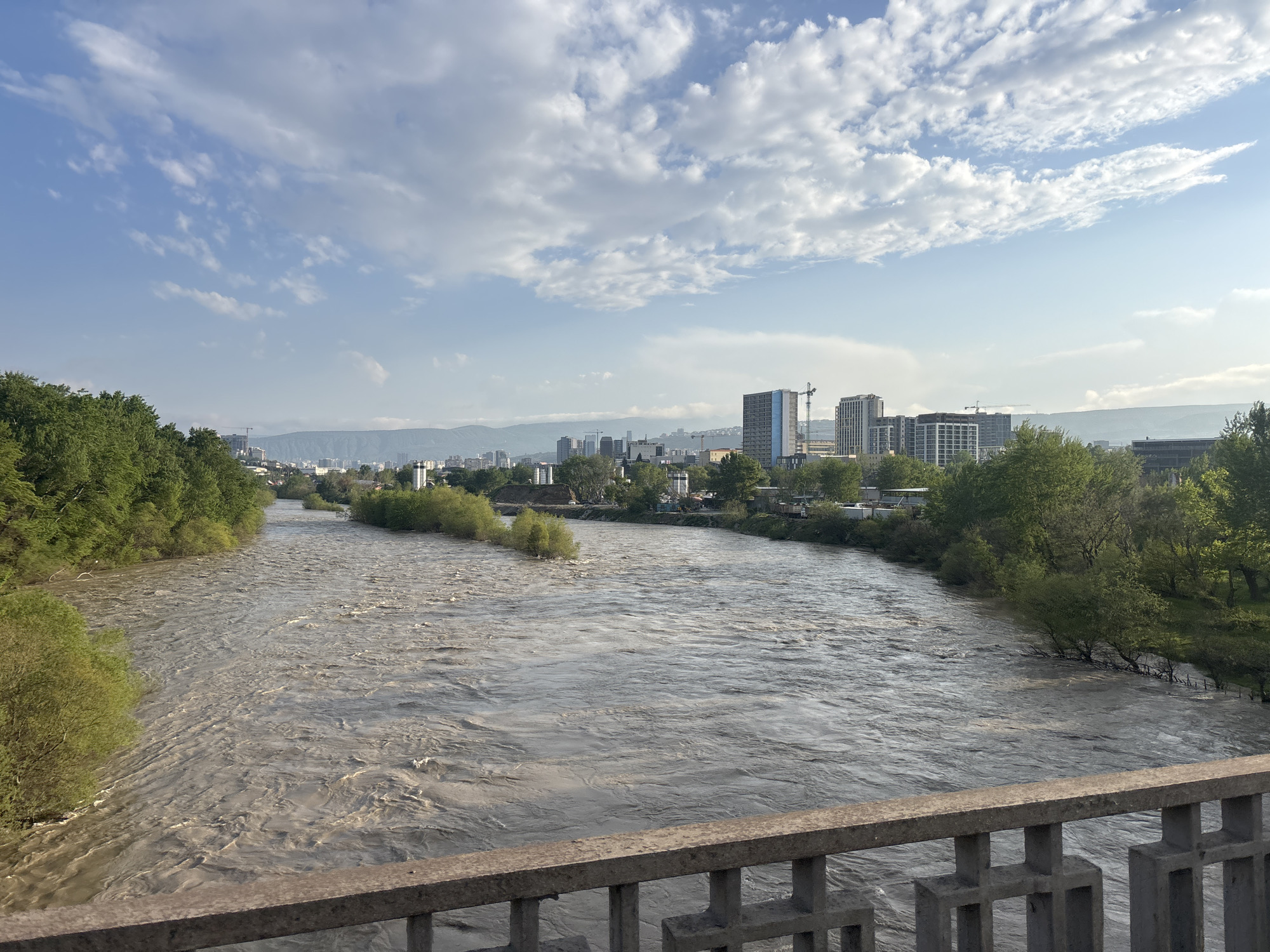 The image depicts a wide, muddy river flowing through Tbilisi, Georgia. The river is flanked by green trees and vegetation on both sides. In the distance, a city skyline is visible, featuring modern buildings and construction cranes. The sky is blue with scattered white clouds. The viewpoint is from a bridge with a gray concrete railing. The water surface is textured with visible currents.