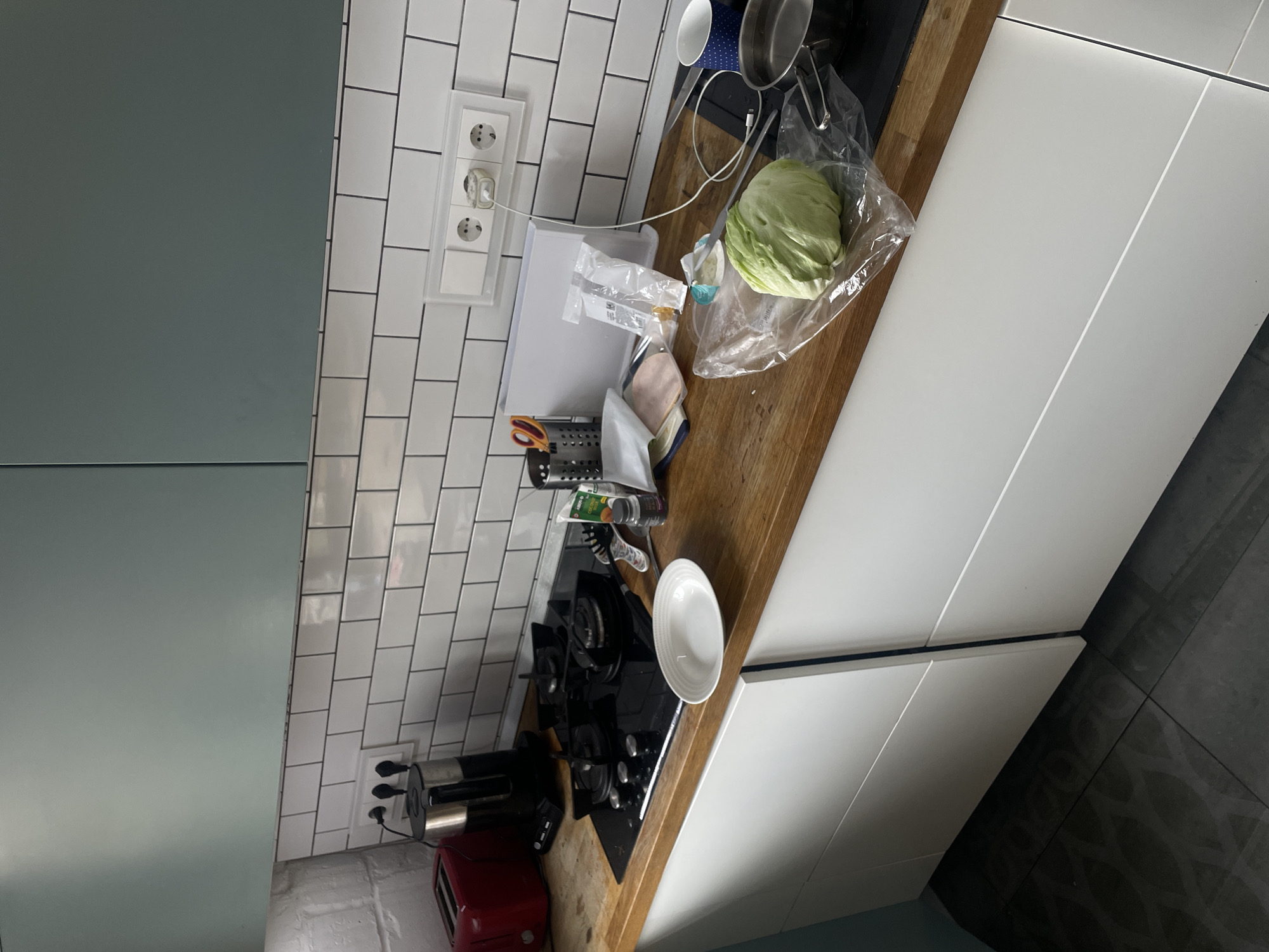 The image presents a section of a kitchen interior. The kitchen features white subway tile backsplash and modern cabinetry with a combination of light gray and white cabinets. On the countertop, there is a black cooktop stove, a red toaster, a white plate, a container of kitchen utensils, food items like a head of cabbage inside a plastic bag, sliced meat, and other packaged goods. There are also cooking pots and mugs present. An electrical outlet is visible on the wall above the countertop, with a charging cable plugged in. The countertop is wooden. The environment appears to be a residential kitchen setting. It is not possible to determine the location as Moscow, Russia without further information or context.