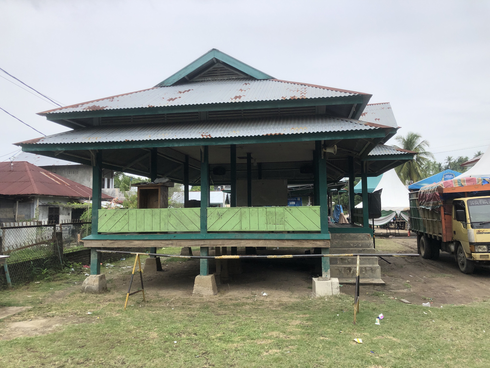 In Palohgadeng, Indonesia, an outdoor stage structure with a multi-tiered, corrugated metal roof, showing signs of rust and weathering, is visible. The stage is painted green and elevated on stilts. Steps lead up to the stage from the right side, adjacent to a dirt path. A yellow truck with a covered cargo bed is parked on the right, near the steps. White tents and palm trees are present in the background, suggesting an outdoor event or market. The ground is covered in grass and dirt, with scattered debris. A chain-link fence with a gate is visible to the left of the stage, bordering a residential building. The sky is overcast. No people are clearly visible on or near the stage.