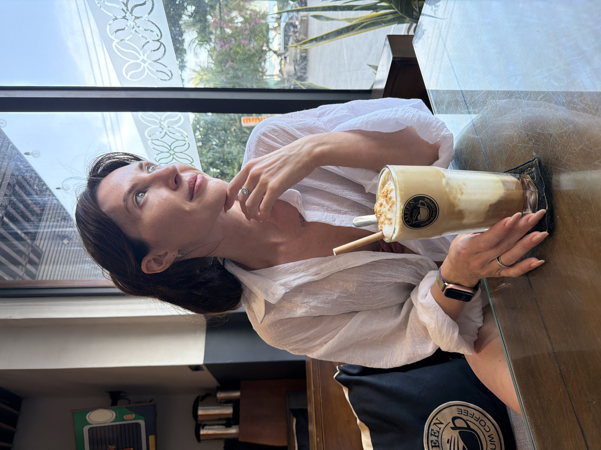 A woman is seated at a glass-topped table inside a cafe, gazing out a large window to her right. She is wearing a loose-fitting white long-sleeved shirt and has dark hair, a watch with a rose gold case and green band on her left wrist, and a silver ring on her left ring finger. Her left hand rests near her chin, while her right hand holds a tall glass of layered iced coffee with cream and toppings, featuring a bamboo straw and a "CALEEN PREMIUM COFFEE" logo. The coffee glass sits on a dark coaster. Behind her to the left, dark shelves display metallic containers and a green decorative sign resembling a vehicle grille. To her left, a black cushion with a matching "CALEEN PREMIUM COFFEE" logo is visible.
Through the window, frosted patterns are visible on the glass. Beyond, a bright blue sky with scattered clouds, lush green trees, and flowering plants create an outdoor scene. A road with parked vehicles, including motorcycles, and what appears to be a distant beach or coastline are discernible, indicating the location is Bac Nha Trang Ward, Vietnam. A potted snake plant is visible in the foreground near the window.