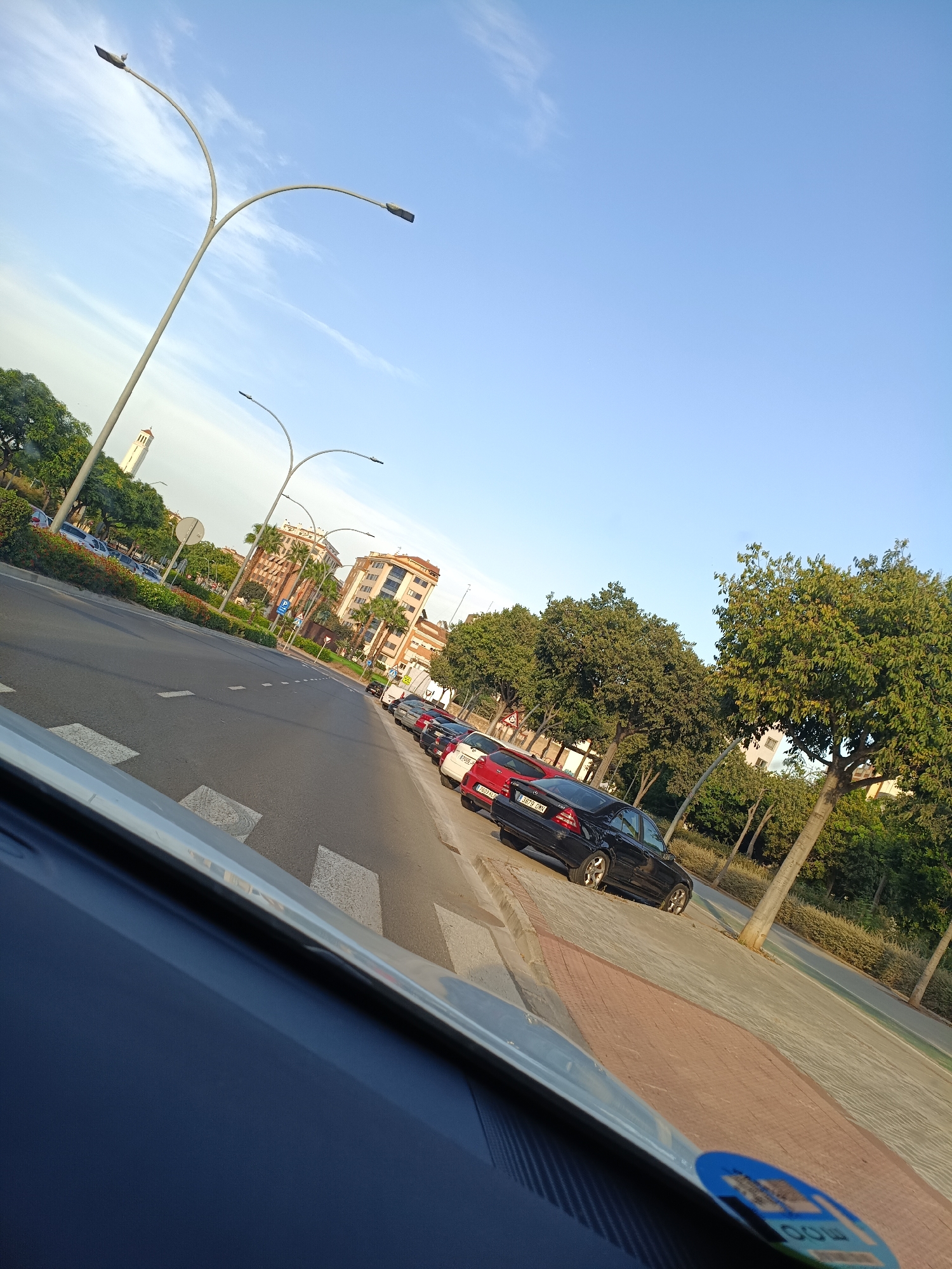 This is a perspective shot taken from inside a vehicle, looking out at a street scene in Castelló de la Plana, Spain. The foreground is dominated by the interior of the car, with the blue dashboard and a silver trim piece visible. Beyond the car, a road with white lane markings and a pedestrian crossing is seen. Several cars are parked along the right side of the street, including a black Mercedes-Benz sedan and a red car. The street is lined with trees and low-lying bushes. In the background, there are multi-story buildings and a distinctive tall, white tower with a red roof. The sky is clear and blue, with a few wispy clouds. The overall impression is of a quiet, sunny day in an urban environment.