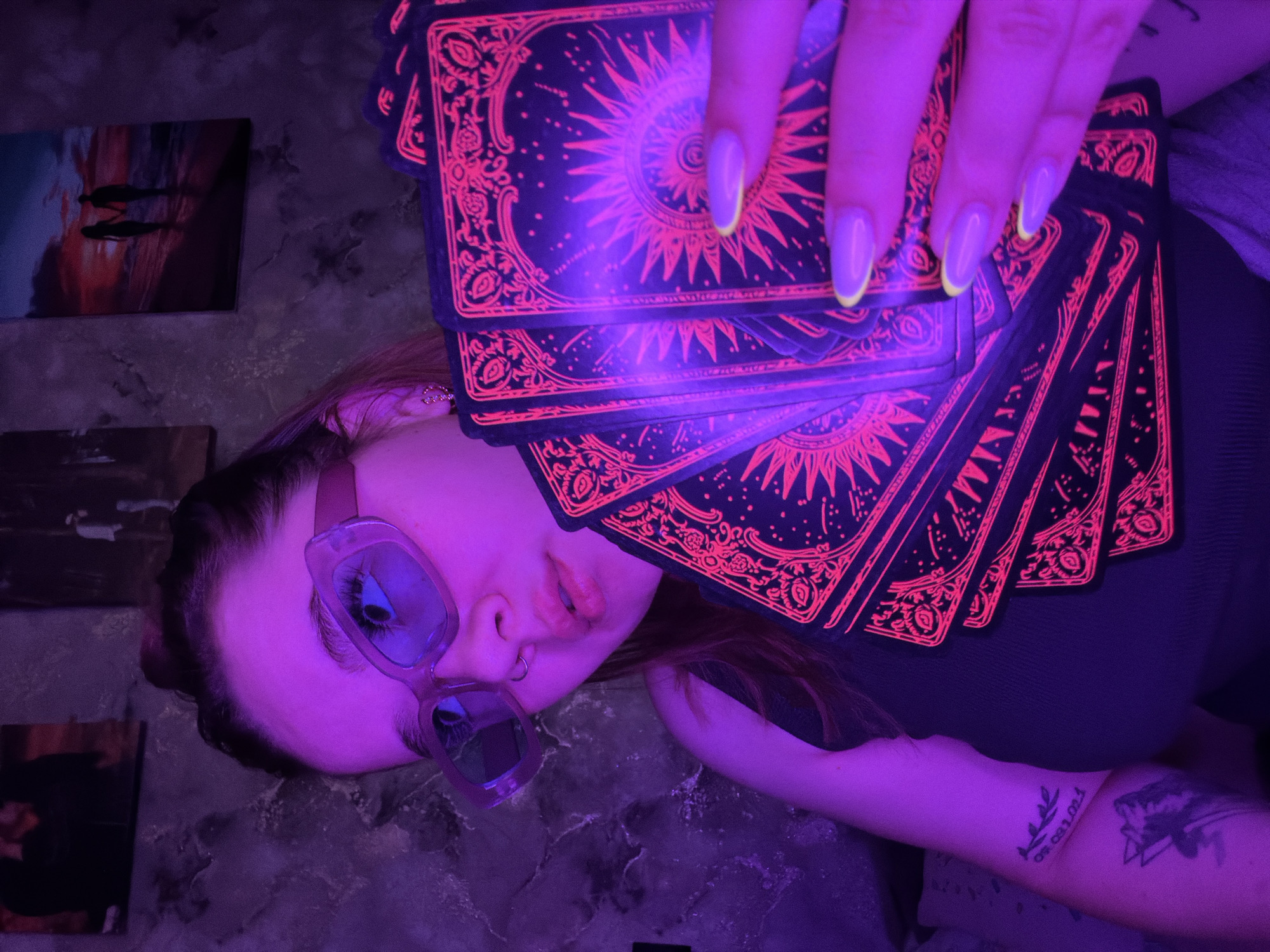 A young adult female is lying on a marble-like surface with her head tilted to the side. She is wearing square-framed sunglasses with reflective lenses, a dark-colored top, and has several tarot cards held in her hand above her body. The cards have intricate designs in red against a dark background. Her nails are long and painted with a light color and a neon yellow edge. A nose ring is visible. She has a tattoo on her left forearm with numbers "09.03.2024" and another one on the same arm with a black geometric shape, and another one on the same arm. In the background, there are framed pictures leaning against the wall. The scene appears to be illuminated with a purplish or UV light. The location is in the city of Chaikovsky, Russia.