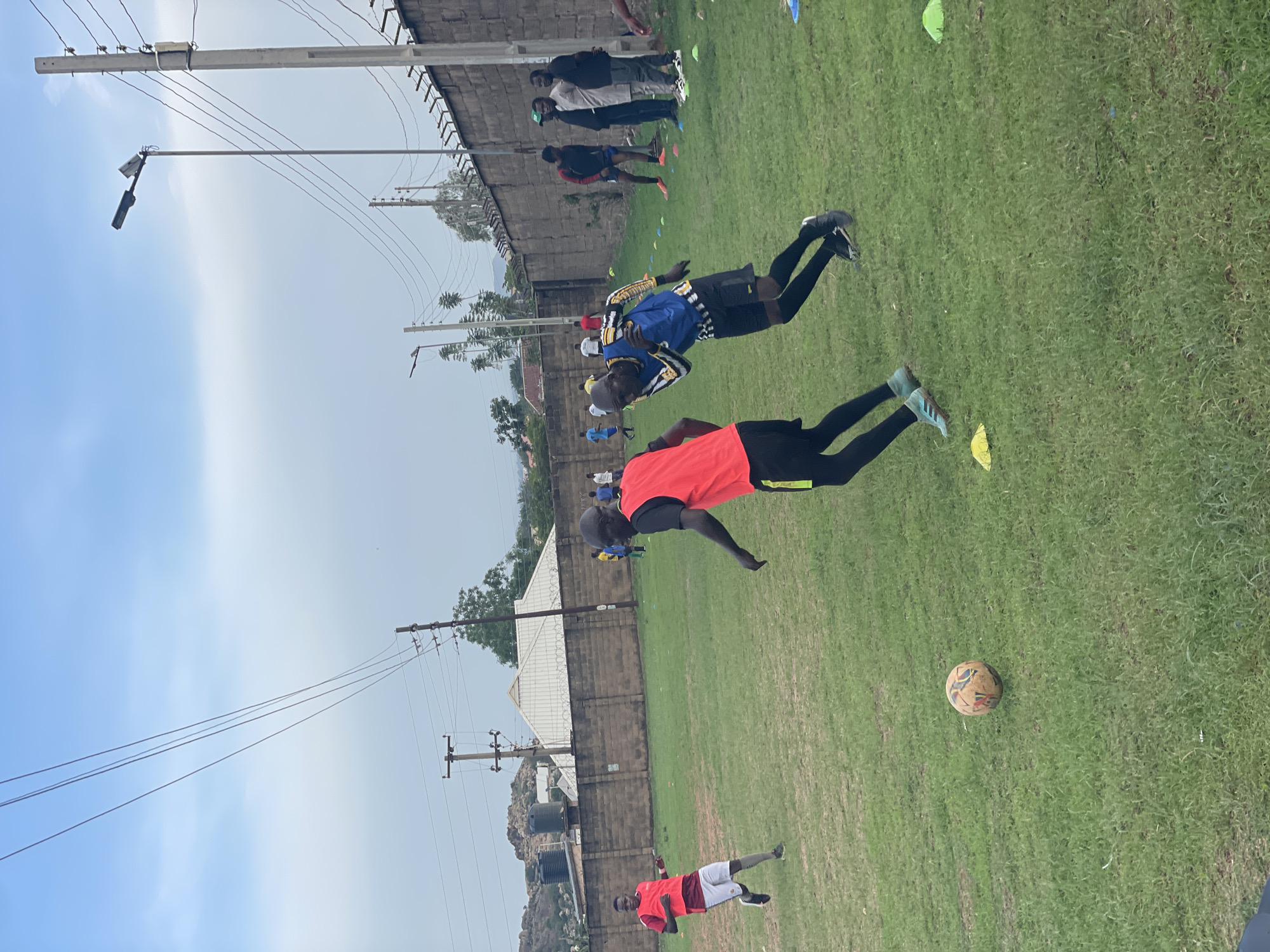 This image, rotated 90 degrees clockwise, captures an outdoor football (soccer) training session or casual game on a grassy field in Jos, Nigeria. The sky is overcast or partly cloudy, indicating daytime conditions with diffused lighting.
Several individuals, predominantly men and youth, are present on the field. In the mid-ground, one person wearing a bright red t-shirt, black leggings, and light blue athletic shoes is shown mid-jump or mid-stride. Close by, another individual in a blue and yellow patterned long-sleeved top, black shorts, black leggings, and black athletic shoes is also in an active pose. A football with visible patterns rests on the green grass. Various small training cones in yellow, blue, and green are dispersed across the field, suggesting organized drills. Toward the lower right, a person in a red t-shirt and white shorts stands on the grass.
In the background, a group of men in casual attire can be seen observing or participating near a long, low wall topped with barbed wire that borders the field. Beyond this wall, buildings with corrugated metal roofs are visible. The scene is also characterized by utility poles carrying overhead power lines and street lights, which extend across the sky. In the distant background, rocky hills and cylindrical black water tanks further define the semi-urban or rural landscape of Jos, Nigeria. No discernible text is visible on any objects or clothing.