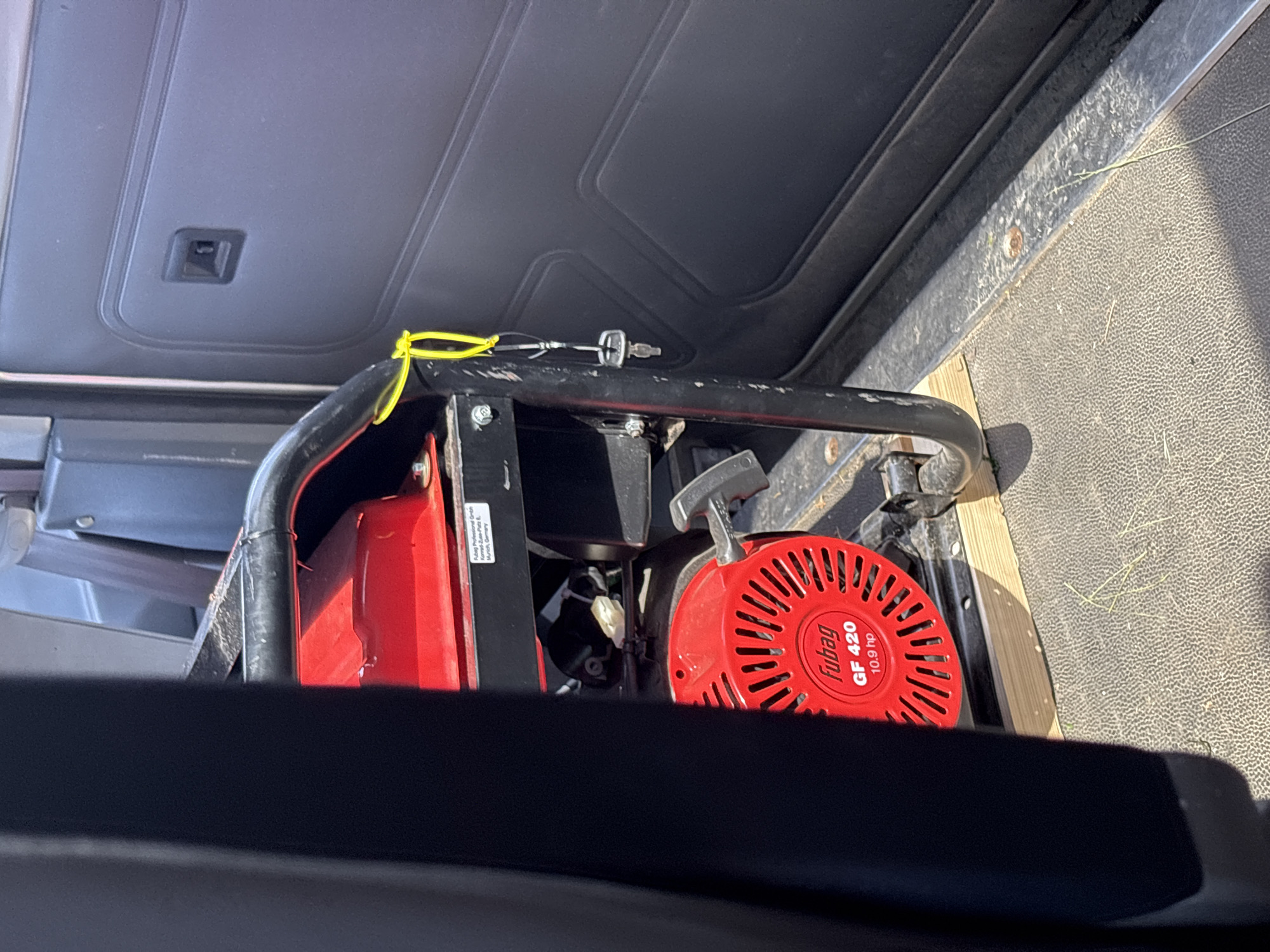 The image shows the interior of a vehicle, likely a utility van or truck, with a portable generator secured inside. The generator is red and black, with the "Fubag GF 420 10.9 hp" branding visible on its red casing. A yellow cord is tied to the generator's frame, and a key is attached to this cord. The generator is resting on a wooden block to stabilize it on the vehicle's floor, which appears to be textured and somewhat worn. The interior of the vehicle shows a dark upholstered panel on the right side, with a recessed handle. The lighting suggests a sunny day, with shadows cast within the vehicle. The general setting suggests a storage or transport context within a rural or suburban area, possibly related to the Мастоцкі сельскі Савет, Belarus, though no specific landmarks or contextual clues point to this precise location within the image itself.