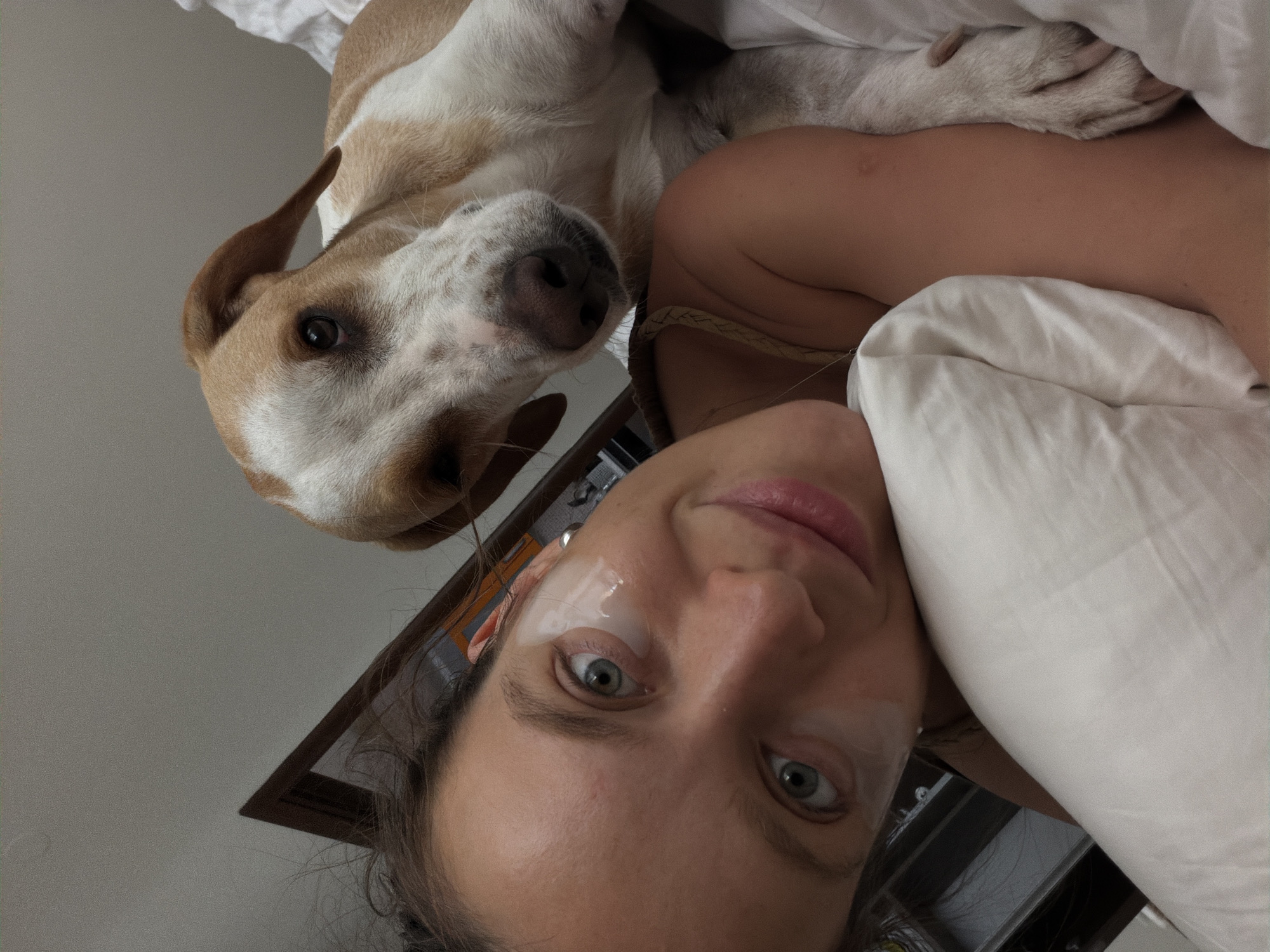 In Budva, Montenegro, a woman with blue eyes and under-eye patches is lying on a pillow in bed, looking at the camera with a slight smile. A light brown and white dog is resting its head on her shoulder, looking towards the camera. The room appears to be a bedroom, with white linens and a wooden headboard visible. The overall impression is an intimate and relaxed moment between a woman and her dog.