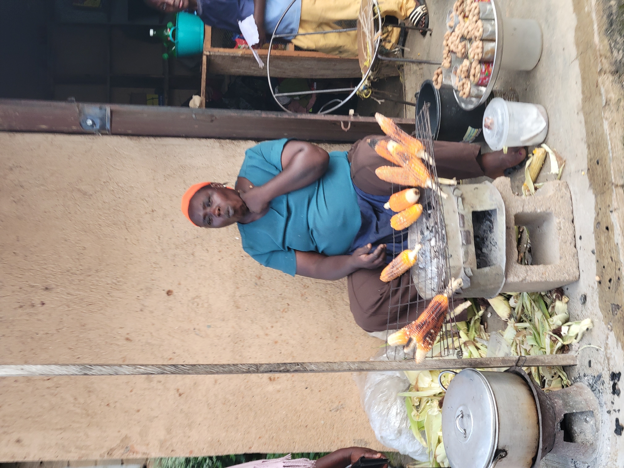 A woman wearing an orange head covering and a teal t-shirt sits in front of a small charcoal grill with several ears of corn cooking on it. She is in the process of eating or preparing to eat one of the corn cobs, with her hand near her mouth. Surrounding her are piles of corn husks, a large metal pot on a stove, and a variety of nuts displayed in small cans on a tray. Another person, partially visible, stands to the right holding some papers. The scene appears to be an outdoor food stall in an urban setting, possibly in a marketplace.