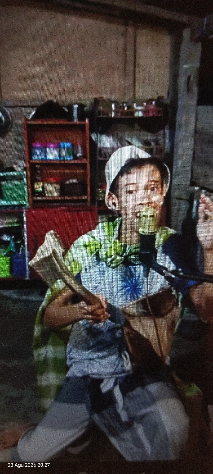 A young male wearing a white cap and a green and white patterned shirt is pictured from the chest up, seated in what appears to be a modest indoor setting. He is holding a rectangular object, possibly a book or a fan, in front of him with both hands, and he is looking directly at the camera with a wide smile. A microphone is positioned in front of his mouth. Behind him, a shelf unit with various items is visible, including what seem to be jars and boxes. The overall lighting suggests an indoor environment, possibly a home or a small studio. The lower-left corner of the image displays a timestamp: "23 Agu 2026 20.27", indicating the date and time the photograph was taken.