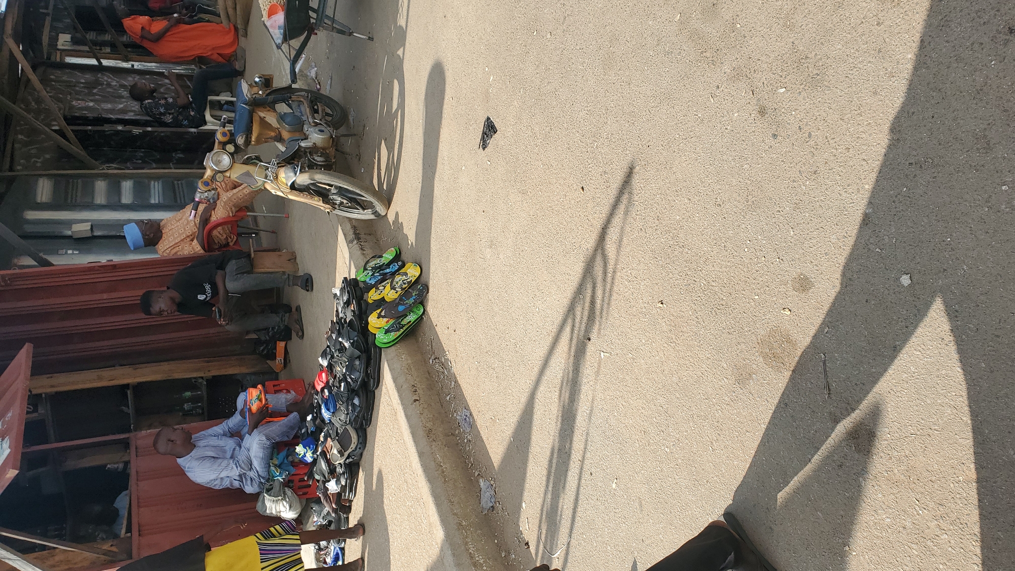 The image depicts a street scene in Jalingo, Nigeria, likely a marketplace. In the foreground, a variety of open-toed footwear, possibly sandals and flip-flops, are displayed on the ground and on a crate. A red crate is visible, holding more footwear. Several individuals are present. On the left, a person wearing a blue and white striped garment sits on the red crate, handling an orange-colored sandal. To their right, a person in dark clothing sits on a wooden stool, looking at their phone. Further to the right, an elderly man in a patterned brown garment and a blue cap is seated on a red chair. Beside him, a yellow motorcycle is parked. In the background, the scene is framed by market stalls with corrugated metal roofing and wooden structures, suggesting a commercial area. The ground is paved with asphalt, and shadows of people and objects are cast across it. Other individuals are visible in the background, including a person in an orange dress. The overall impression is of a bustling, open-air market environment.