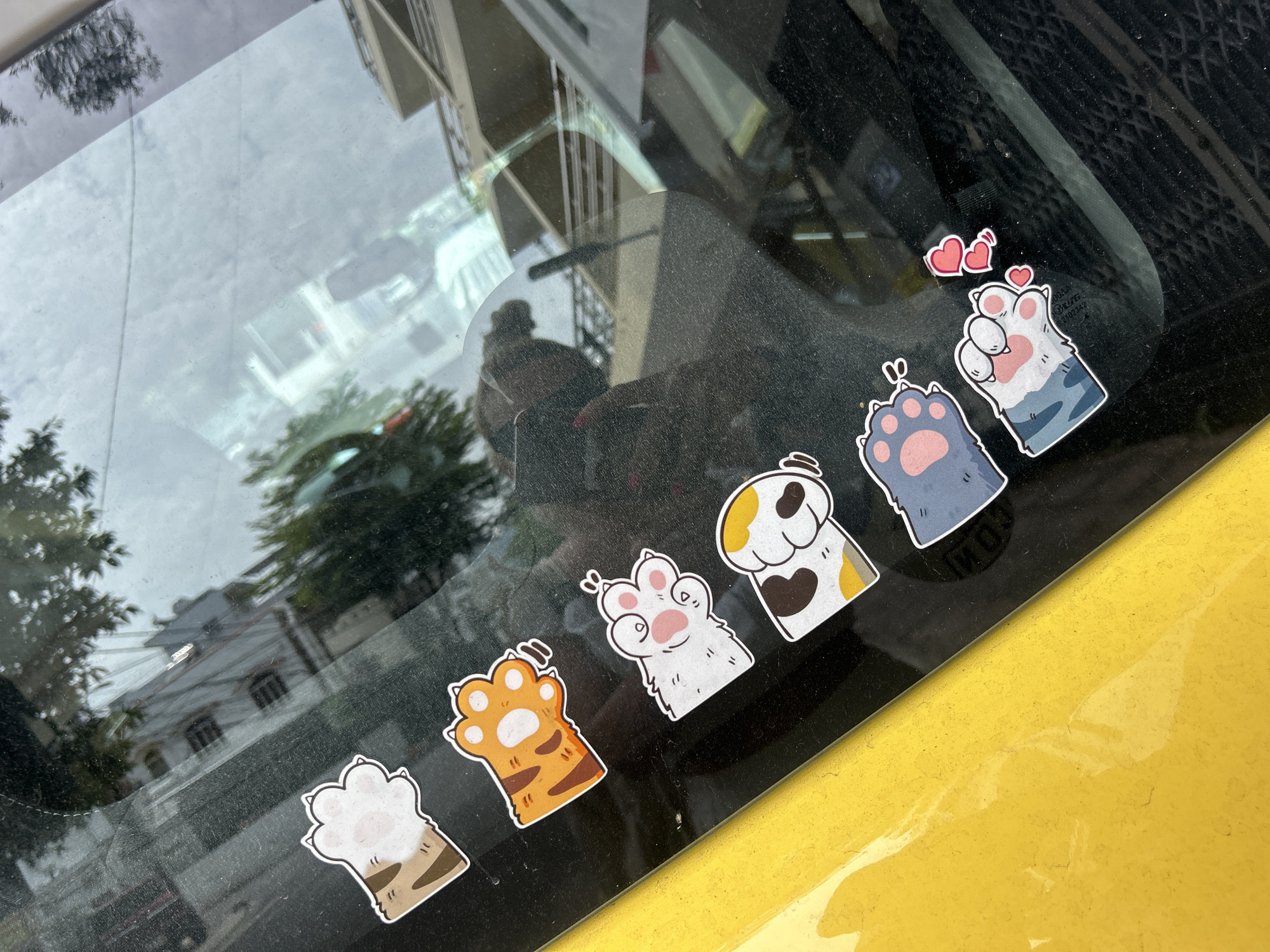 The image shows the interior of a vehicle, specifically the rear window, adorned with several cartoon cat paw stickers. The window is tinted, and the background is partially obscured but appears to be an urban landscape. Buildings and trees are visible through the window. The car's exterior is yellow. The location may be in or around Bac Nha Trang Ward, Vietnam, as it is mentioned in the prompt.