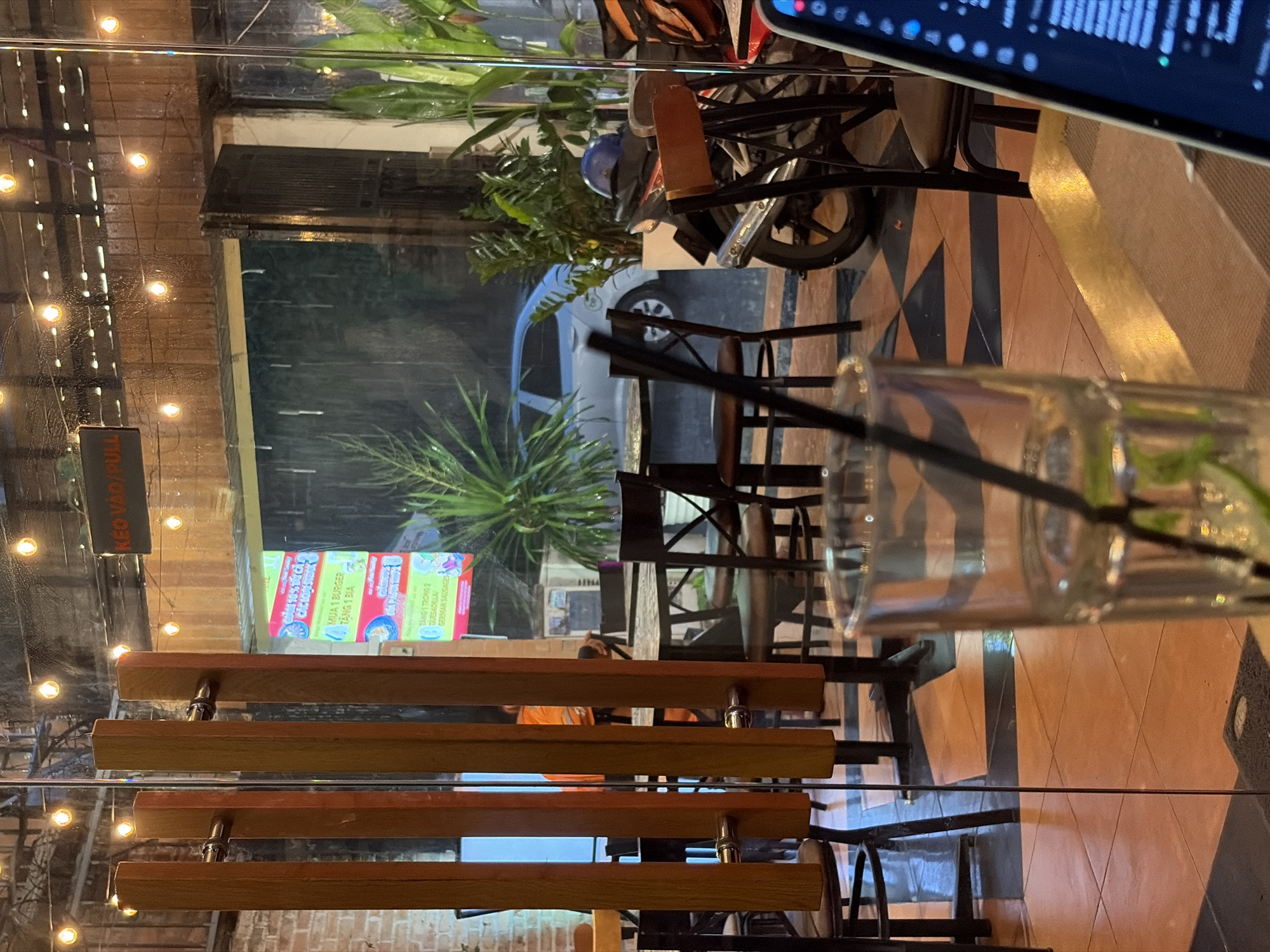 The image depicts an interior view of a restaurant in Thủ Đức, Vietnam, on a rainy night. Through a glass partition with wooden vertical elements, a dimly lit outdoor area is visible. Rain streaks are apparent on the glass. The outdoor space features a parked motorcycle with a blue helmet on it, a light-colored car, and lush green plants. Inside the restaurant, several tables and chairs are set up. A partially visible glass of water with a straw and lime slices is in the foreground, resting on a table next to an open laptop. The floor has a distinctive orange and black tiled pattern. A sign with Vietnamese text, indicating "PULL," is mounted above the entrance. The overall atmosphere suggests a cozy, possibly wet, evening dining experience.
