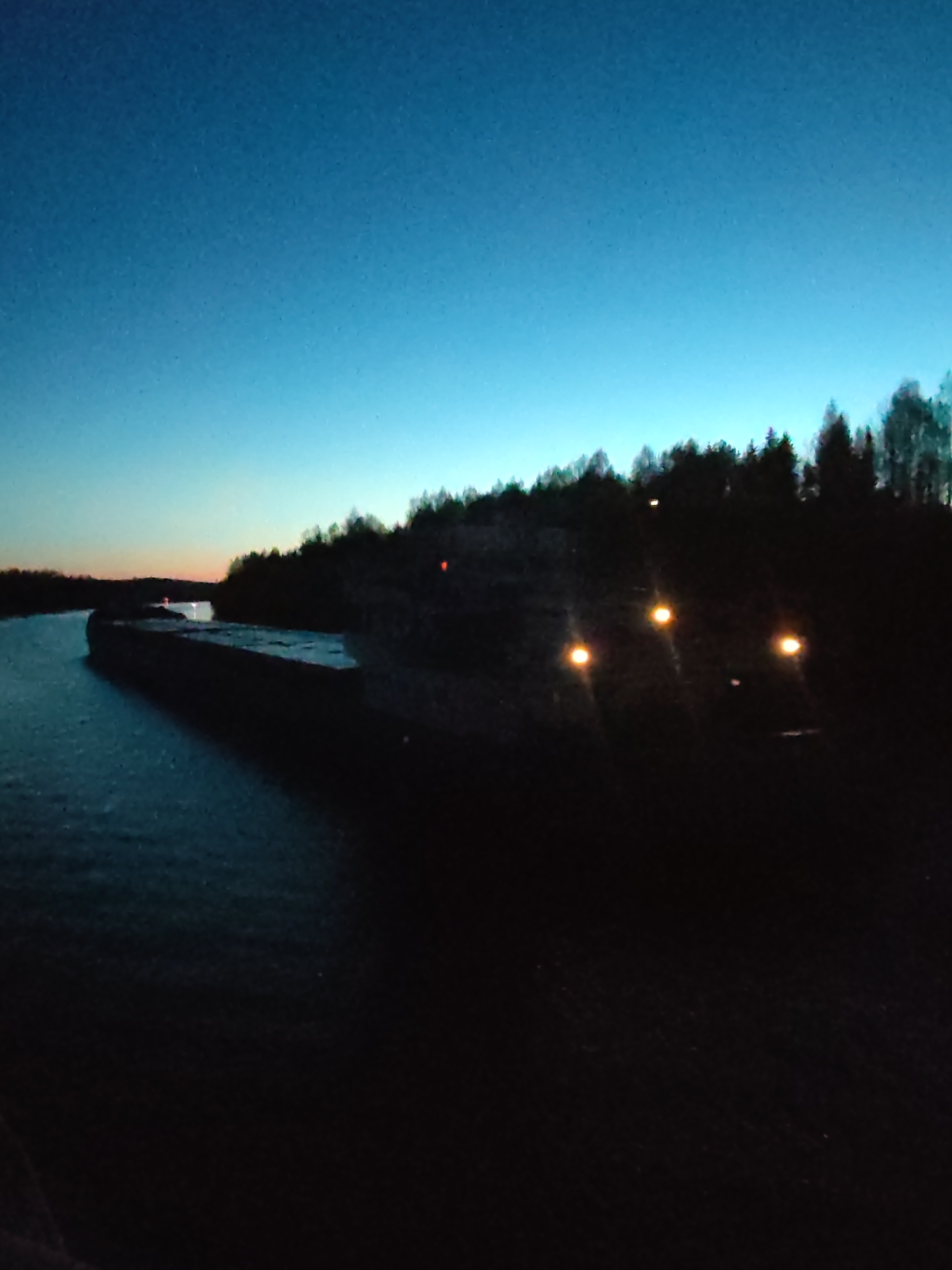The image depicts a waterfront scene in Uzhla, Russia, at dusk or twilight. The sky is a gradient of blue, fading into orange near the horizon. A body of water, possibly a river, occupies the left side of the frame, with a dark structure or embankment running along its edge. Across the water is a treeline silhouetted against the sky. Further back from the embankment are a few dim lights, with three bright lights illuminating an area to the right of the embankment. The foreground is dark and mostly obscured.