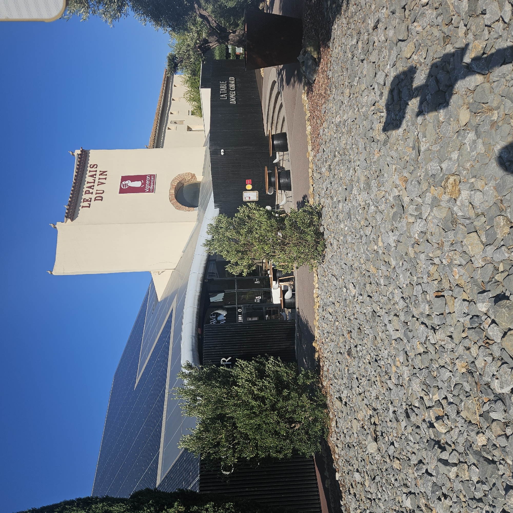 The image shows the exterior of "Le Palais du Vin" in Narbonne, France. The building features a large section of solar panels on its roof and a cream-colored tower with the text "LE PALAIS DU VIN" in dark red lettering. Below this text is a logo for "vigneron indépendant". The building has a modern architectural style with dark vertical siding on the lower sections.
In front of the building, there are outdoor seating areas with wooden tables and barrel-like bases, and chairs. A large potted olive tree stands to the right of the entrance. The ground in the foreground is covered with grey and brown gravel and stones. The sky is clear and blue. Shadows of people are visible on the ground in the lower right portion of the image, suggesting the presence of visitors. The overall scene depicts a commercial establishment focused on wine, with a contemporary and eco-friendly design.