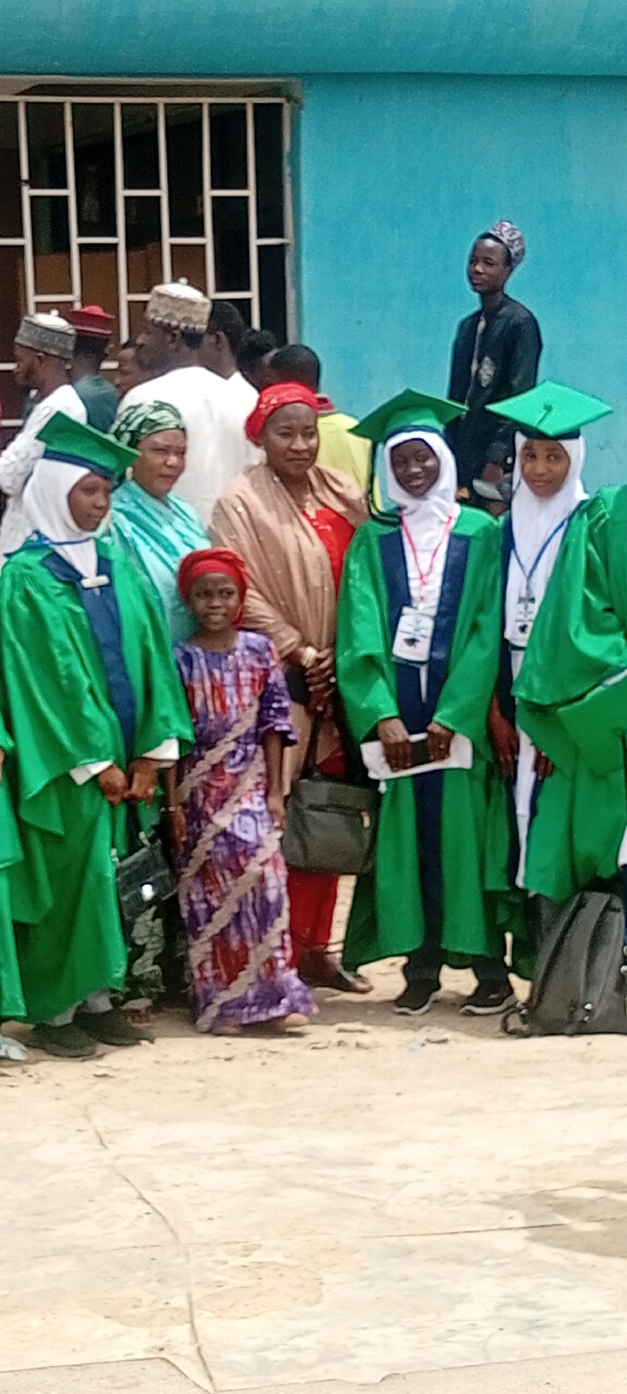 The image depicts a group of people, primarily women and children, some of whom are wearing green graduation gowns and mortarboards, suggesting an academic ceremony or graduation. Several adults are present, and there are men in traditional Muslim attire in the background. They are standing outdoors on a paved surface, and behind them is a light blue building with a barred window. The scene appears to be a public gathering, possibly a graduation celebration. Sabon Gari East, Nigeria is mentioned, but the location is difficult to establish from this view alone.