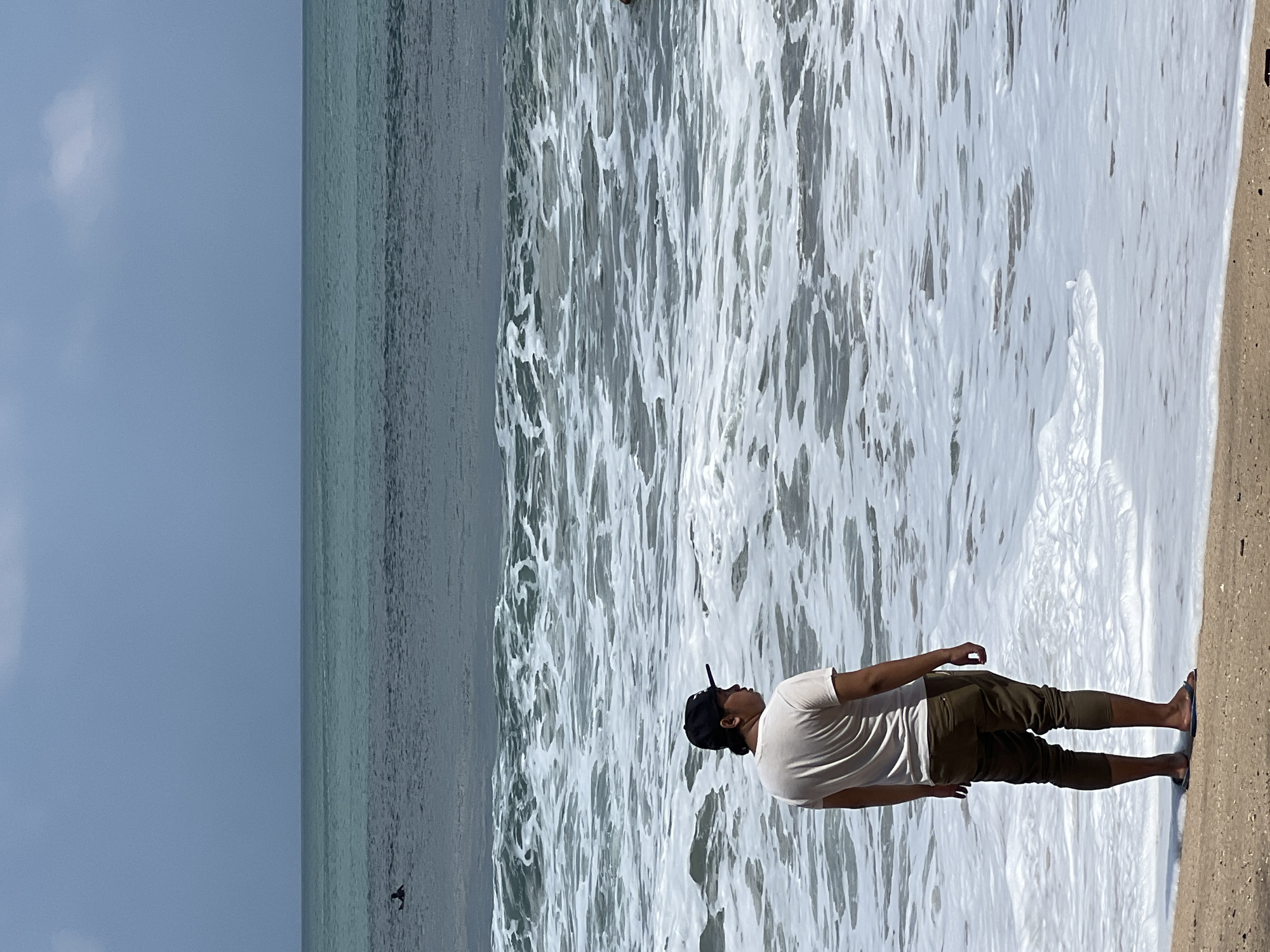 A person stands on the beach at Kuta, Indonesia, facing the ocean. The person is wearing a white t-shirt, olive-colored pants, and a black baseball cap. They are barefoot, with one foot in the shallow water. The ocean is a deep teal color with white foam from waves washing ashore. In the distance, the horizon is visible, separating the water from a pale blue sky with a few wispy clouds. There are small white birds wading in the shallow water.