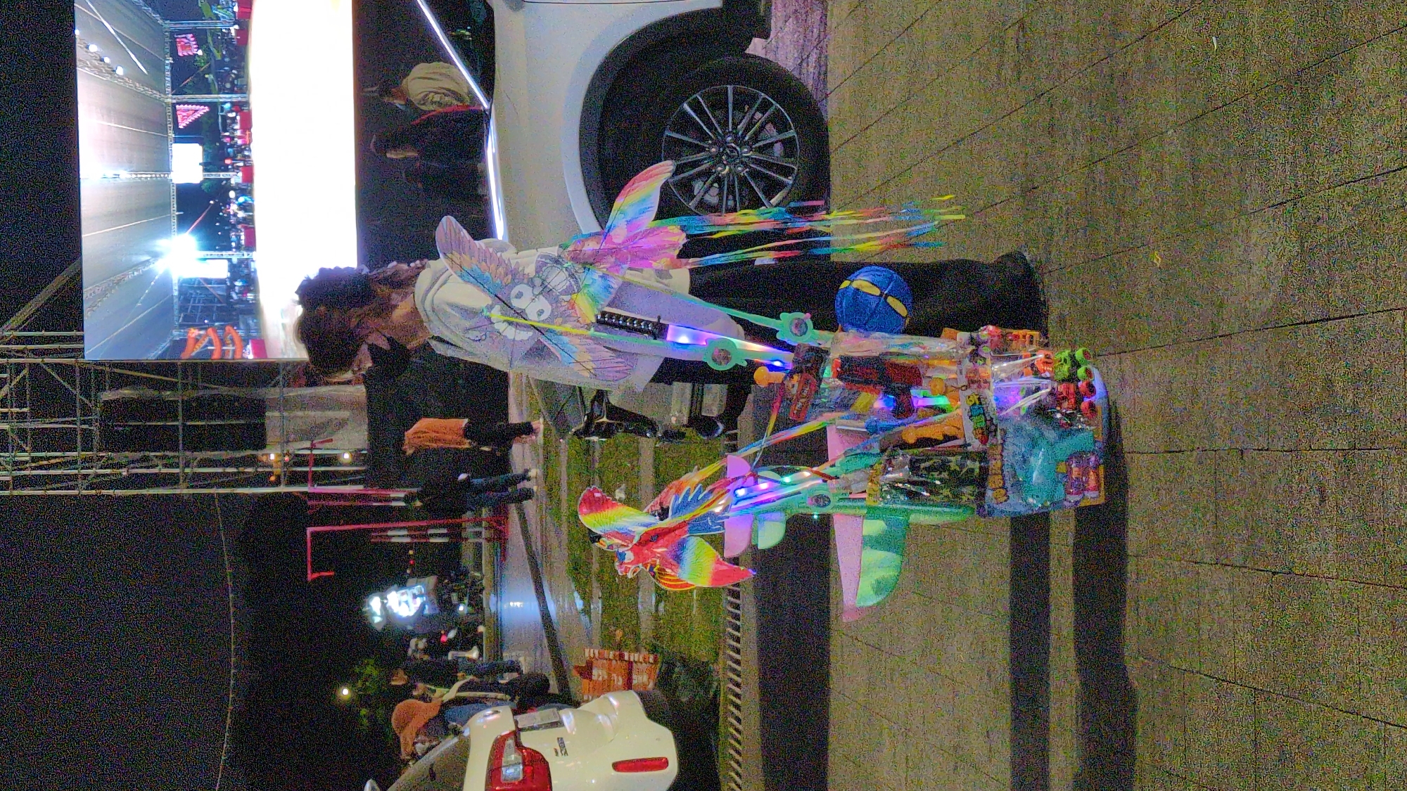 A person with dark hair and a mask stands on a paved surface, displaying a variety of toys, including light-up wings and toy guns. These items appear to be for sale. A white car is parked nearby. In the background, a screen shows a race track. There are additional people present in the scene, as well as cars. The location is likely Da Lat, Vietnam, based on the visual style and context of the image.