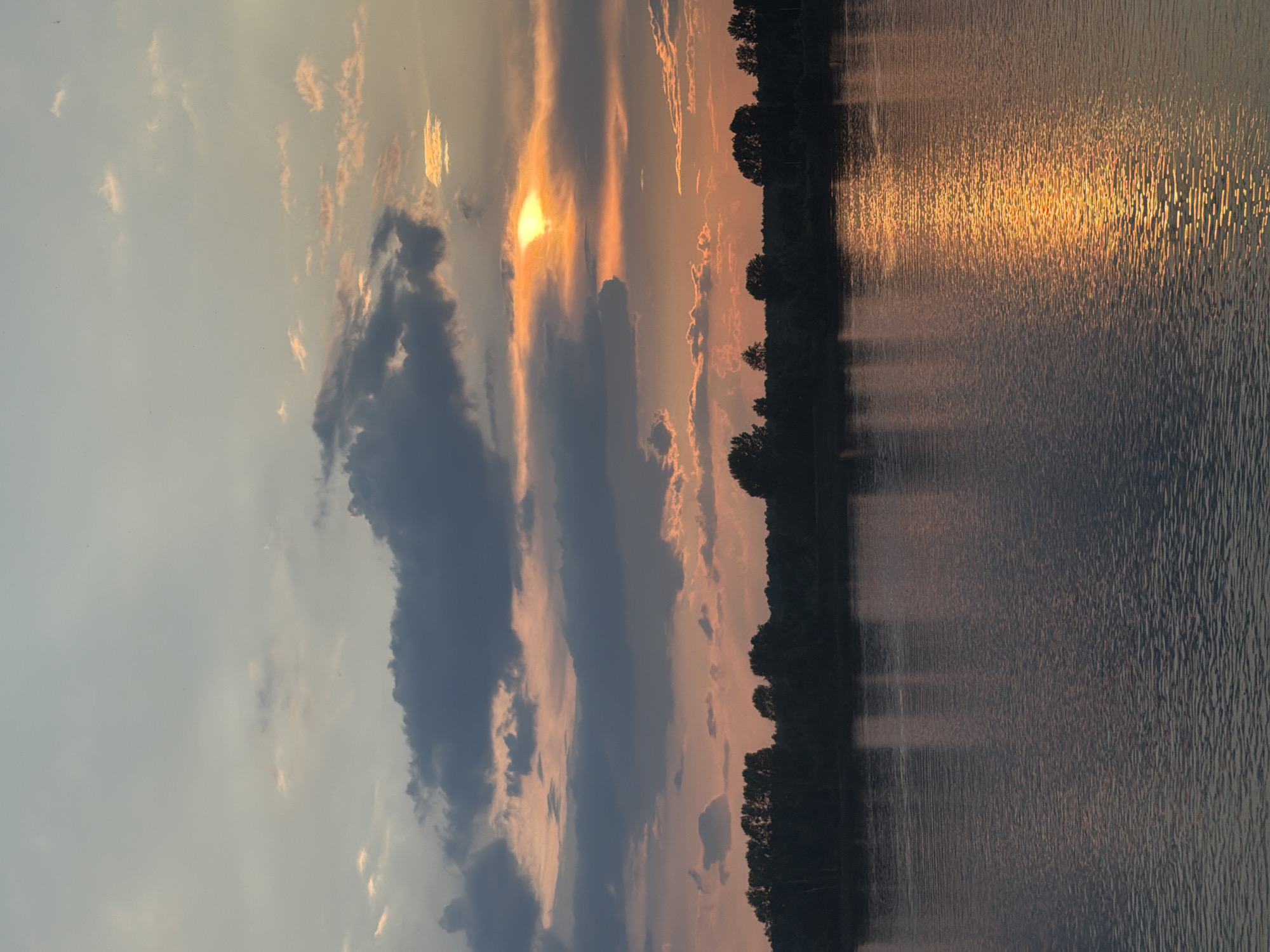 The image depicts a sunset over the Kama River in Chaikovsky, Russia. The sky is filled with a mixture of dark and light clouds, with the sun peeking through, casting a golden and orange glow on the horizon. A line of trees forms the silhouette of the distant shore. The river's surface reflects the colors of the sky, with a bright streak of sunlight stretching across the water. There are no people or any other discernible subjects in the image. The overall scene is serene and natural.