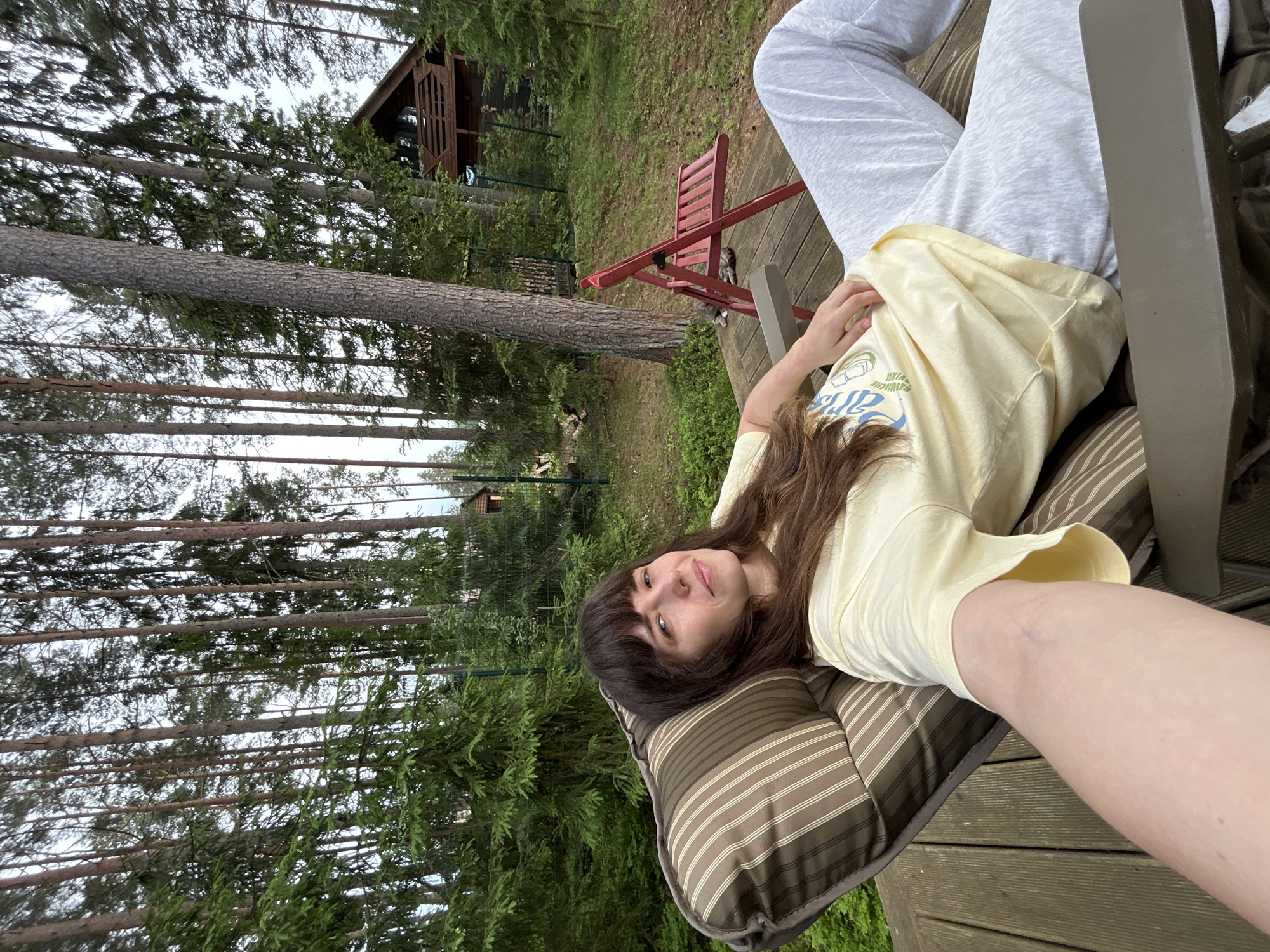 In Shepelyovo, Russia, a woman with long, dark brown hair and bangs is relaxing on a striped cushion chair on a wooden deck. She is wearing a light yellow t-shirt with a graphic design and grey sweatpants. Her right arm is extended forward, resting on the chair. In the background, a dense forest of tall pine trees is visible, with a rustic wooden cabin situated deeper within the woods. A red foldable chair is placed on the deck near the trees, and two cats are lying on the deck in front of it. The overall scene suggests a peaceful, outdoor setting.