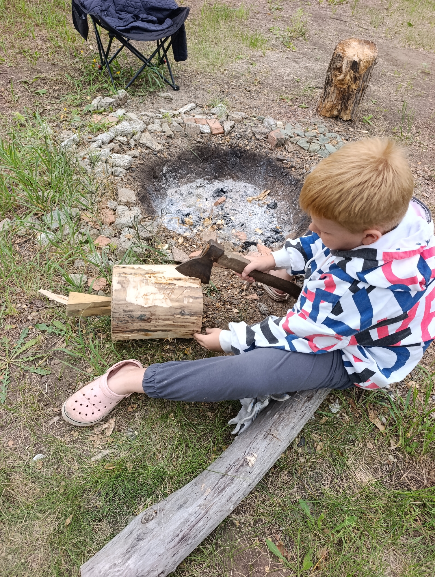 In Dimitrovskii, Russia, a young child with blond hair is sitting on the ground near a fire pit. The child is wearing a hooded jacket with a colorful geometric pattern, grey pants, and pink clogs. The child is holding an axe and appears to be attempting to chop a piece of wood that is propped up on smaller pieces of wood. The fire pit contains grey ash and embers. In the background, there is a black folding chair and a log. The ground is covered in dry grass and dirt, with scattered rocks and small pieces of wood.