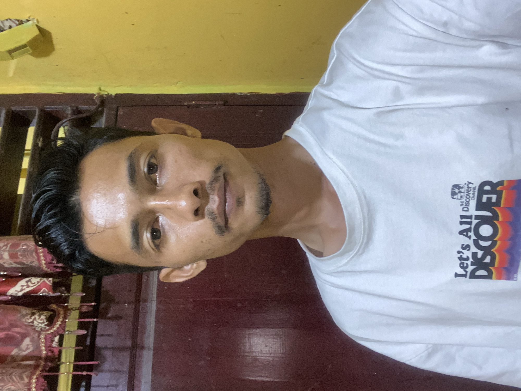 A man with dark hair and a light-skinned complexion poses for a photo indoors. He is wearing a white t-shirt with the words "Let's All Discover" and the Discovery Channel logo printed on it. The background consists of a dark red-brown door or wall, with a yellow wall visible above. To the left, there is a decorative element with red and gold detailing. There is no information available about the location in the city of Tanjongkapai, Indonesia.