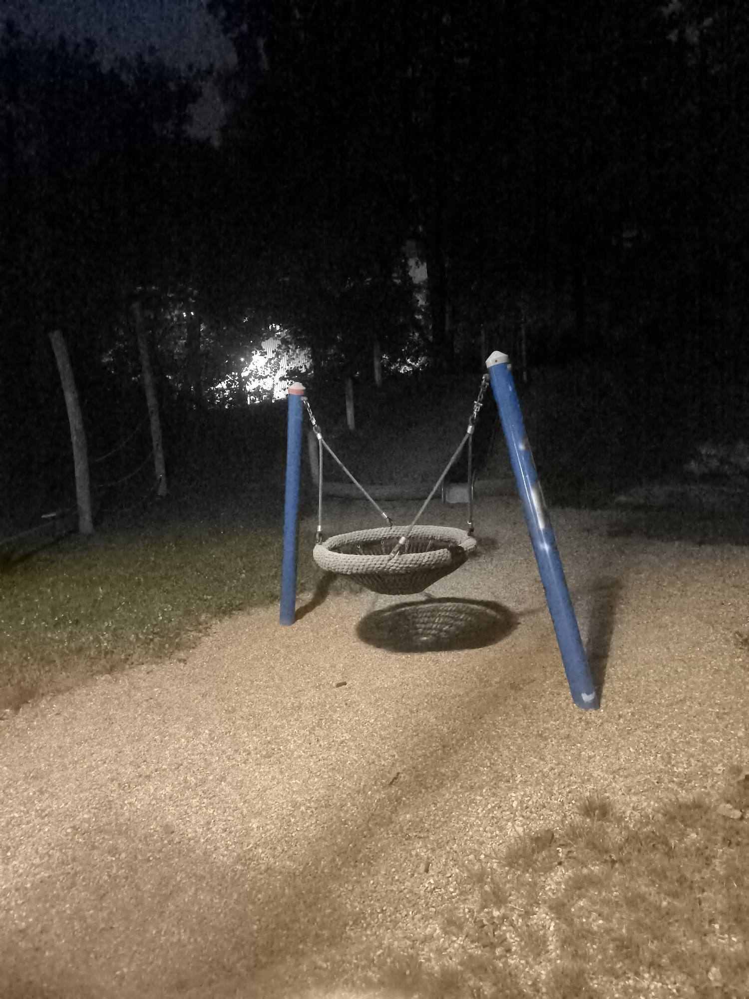 An outdoor, night-time photograph depicts a basket swing in an empty park. The swing is made of woven rope and hangs from two blue poles that are connected by a metal bar. The swing is positioned on a surface of light brown gravel, with a patch of green grass to the left. In the background, there are several trees and some fence posts. The overall lighting is dim, suggesting it is either late at night or the area is not well-lit. No people are present in the image.
