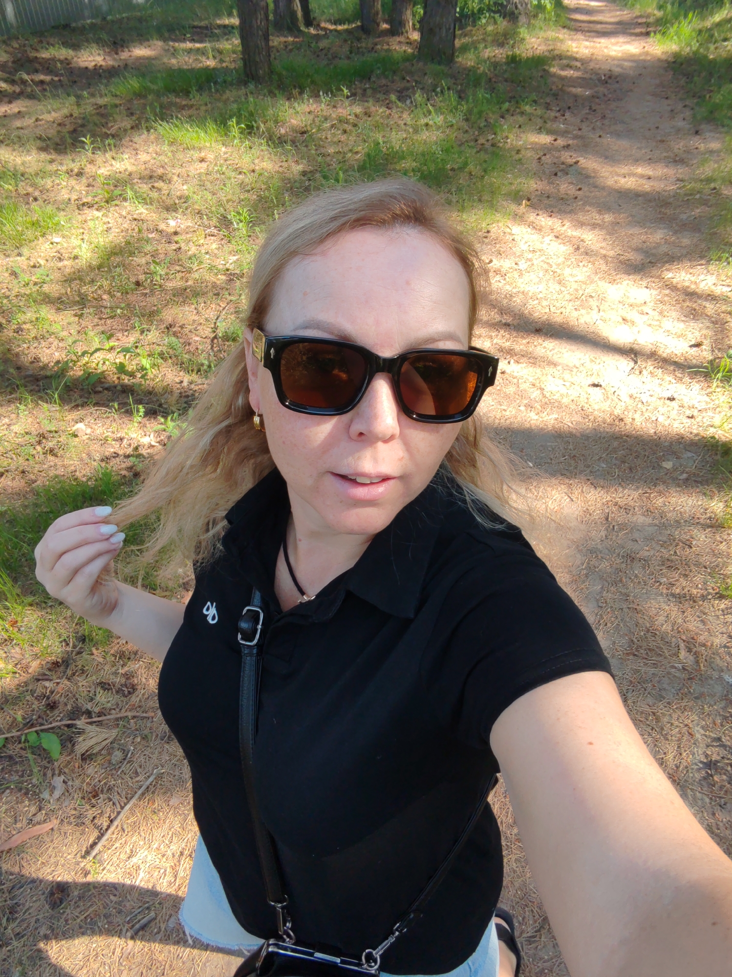 A fair-skinned woman with blond hair and sunglasses is taking a selfie on a dirt path in a wooded area. She is wearing a black polo shirt and a black purse hangs across her body. The location is in Chaikovsky, Russia. Tall trees and dappled sunlight are visible in the background.