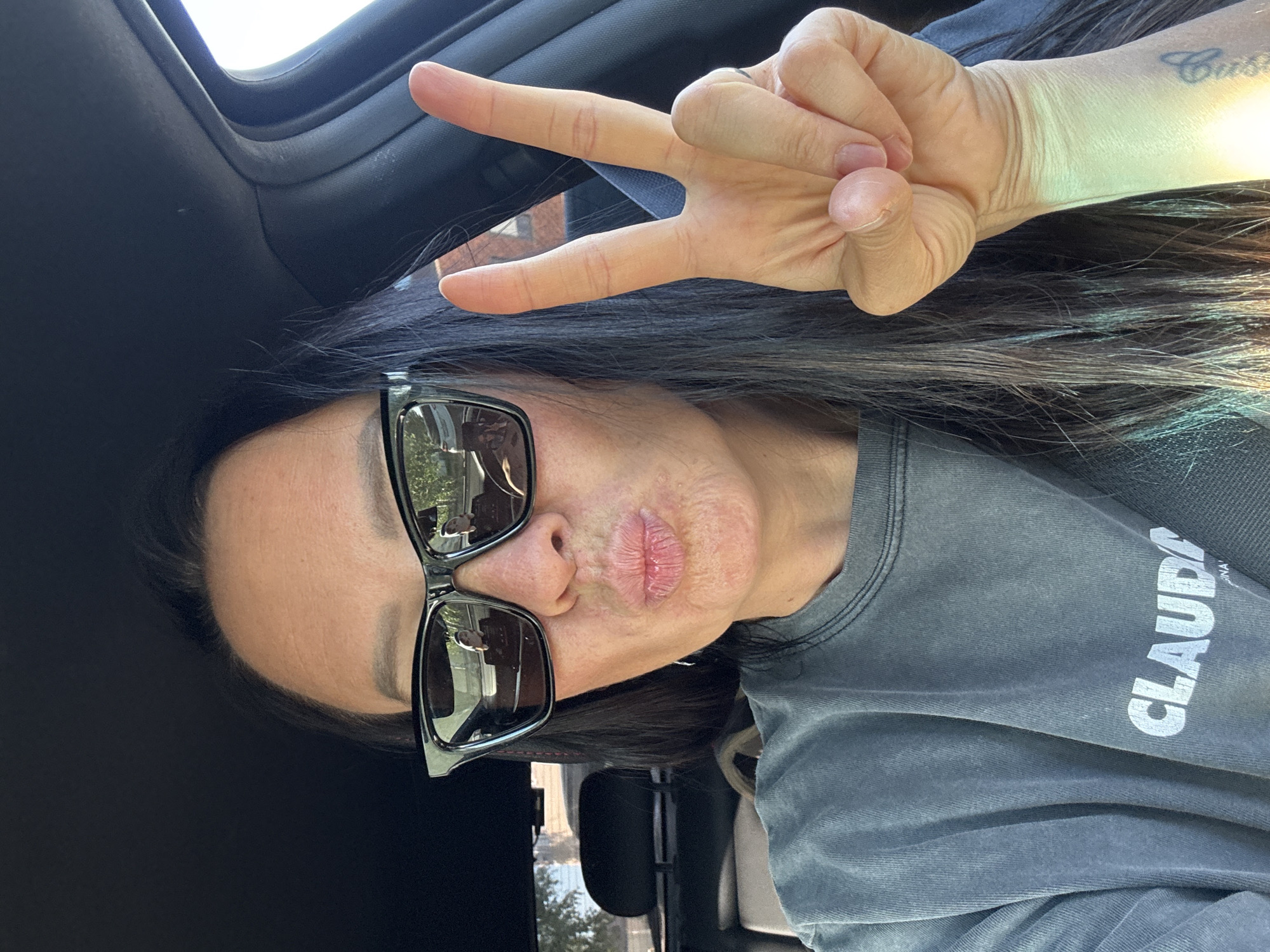A woman is pictured from the chest up, turned to the right, making a peace sign with her right hand. She is wearing sunglasses, a grey t-shirt with the name "Claudia" printed on it. She has dark hair. She is indoors, likely in a vehicle. The location in Chaikovsky, Russia is not discernable from the image.