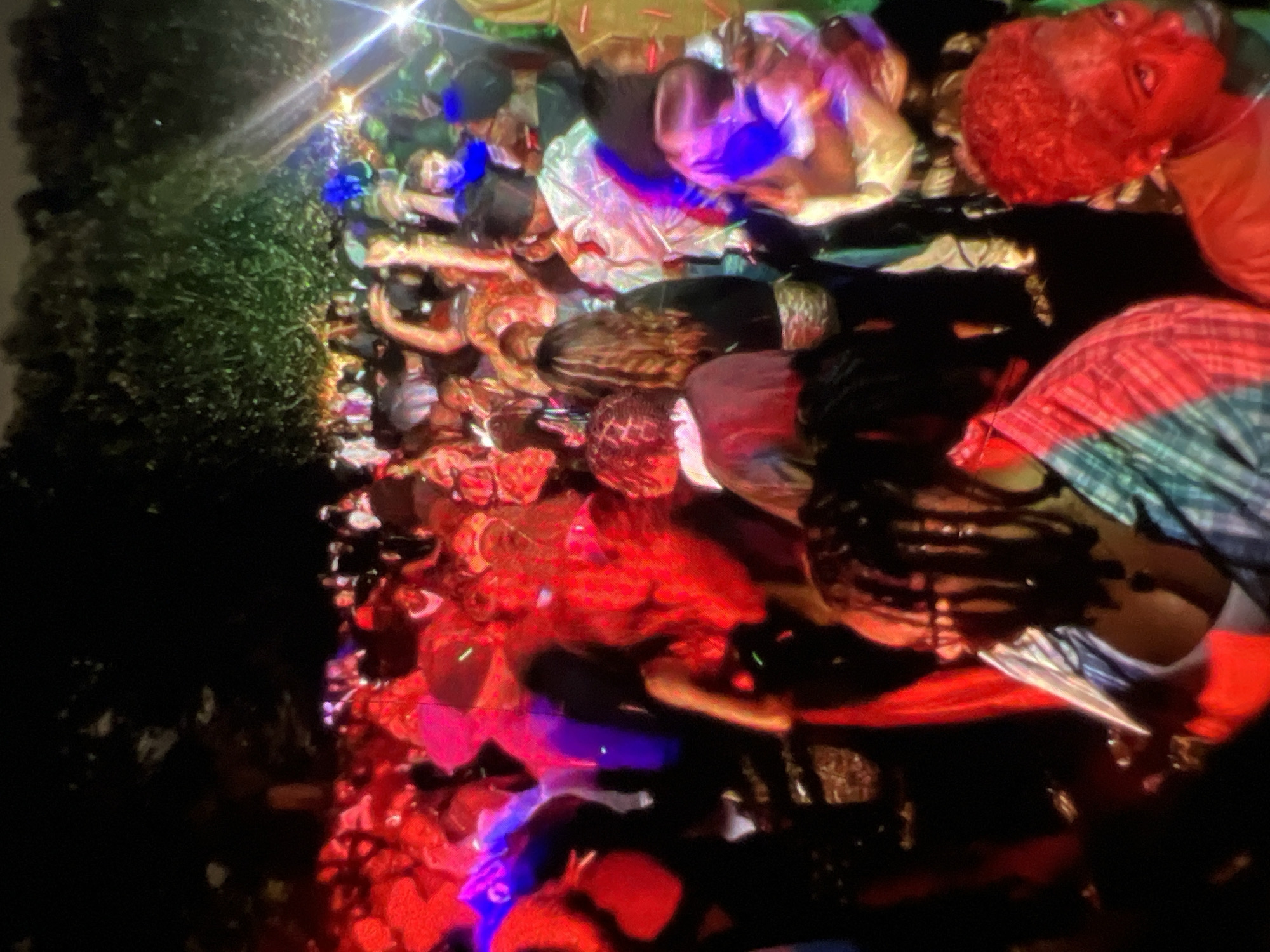 The image depicts a crowded outdoor gathering at night, likely a party or concert, taking place in Lagos, Nigeria. The scene is illuminated by an array of colorful lights, predominantly red and blue, casting vibrant hues across the faces and clothing of the attendees. The background is dominated by dark trees and foliage, suggesting a natural or park-like setting within the city. Many individuals are visible, with some appearing to be dancing or interacting with each other. The overall atmosphere appears lively and energetic, characteristic of nocturnal events in a bustling metropolis like Lagos.