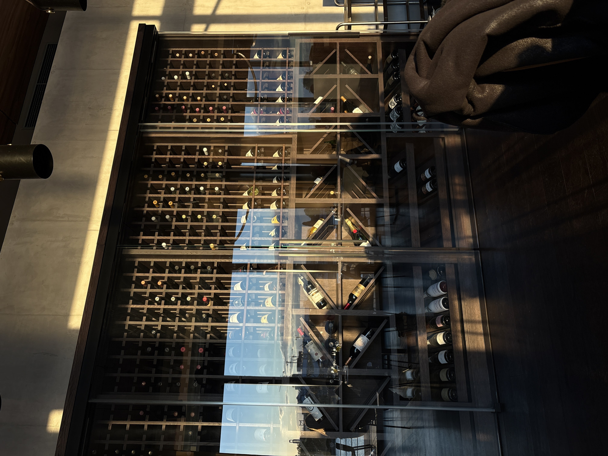 The image shows a wine cellar, with bottles neatly arranged in racks behind glass panels. The location is inferred to be Belgrade, Serbia based on reflected scenery. The cellar contains both horizontally stacked bottles in grid-like sections and bottles stored in individual angular compartments. There is an out-of-focus brown object, possibly furniture, on the right. The floor is dark wood. The scene is well-lit. Reflections of the sky are visible on the glass panels.