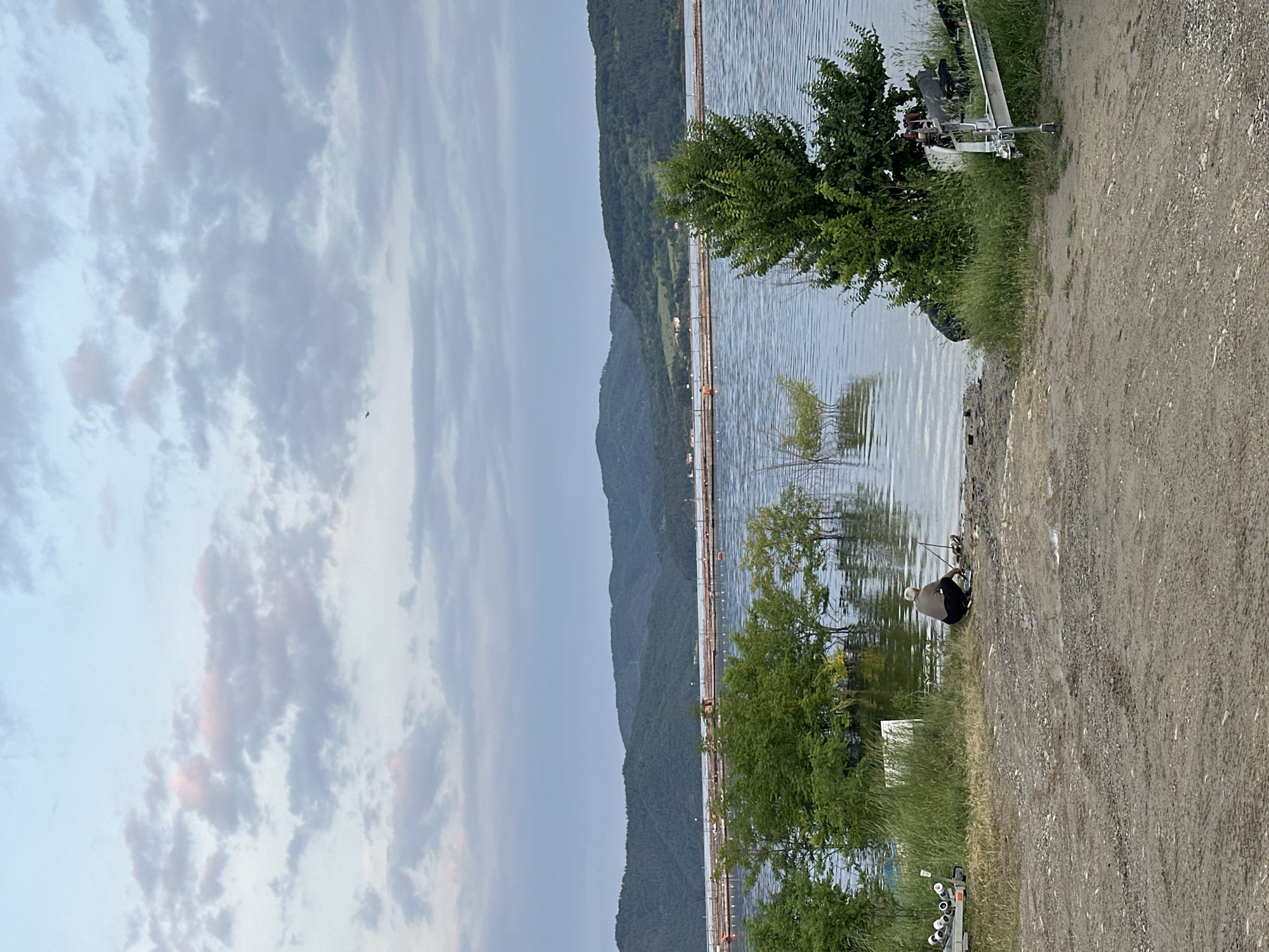This image, oriented 90 degrees clockwise from a natural landscape view, depicts a serene outdoor scene in Brosh, Bulgaria.
On the left side of the frame, an expansive, overcast sky dominates, filled with soft grey and white clouds. Subtle pink and orange hues are visible in some clouds, hinting at either dawn or dusk. A tiny, dark speck, possibly a bird, can be seen flying in the sky.
The middle section features a broad body of water, likely a lake or wide river, with gentle ripples disturbing its surface. Along the far bank of the water, a range of forested mountains rises, stretching across the horizon. A long, linear structure, possibly a series of fishing platforms, nets, or a walkway, spans the water parallel to the distant mountains.
The right side of the image reveals a rugged dirt and gravel path or bank, suggesting a natural or semi-developed shoreline. Green grass and leafy trees/bushes grow sparsely along the water's edge. A lone individual, appearing to be a man, is visible crouching close to the water, engaged in fishing. Several fishing rods are set up around him, indicating active participation in the activity. Various pieces of metallic and white equipment or structures are partially visible further along the bank, near the water.
The overall atmosphere is peaceful and tranquil. No visible text is present in the image.