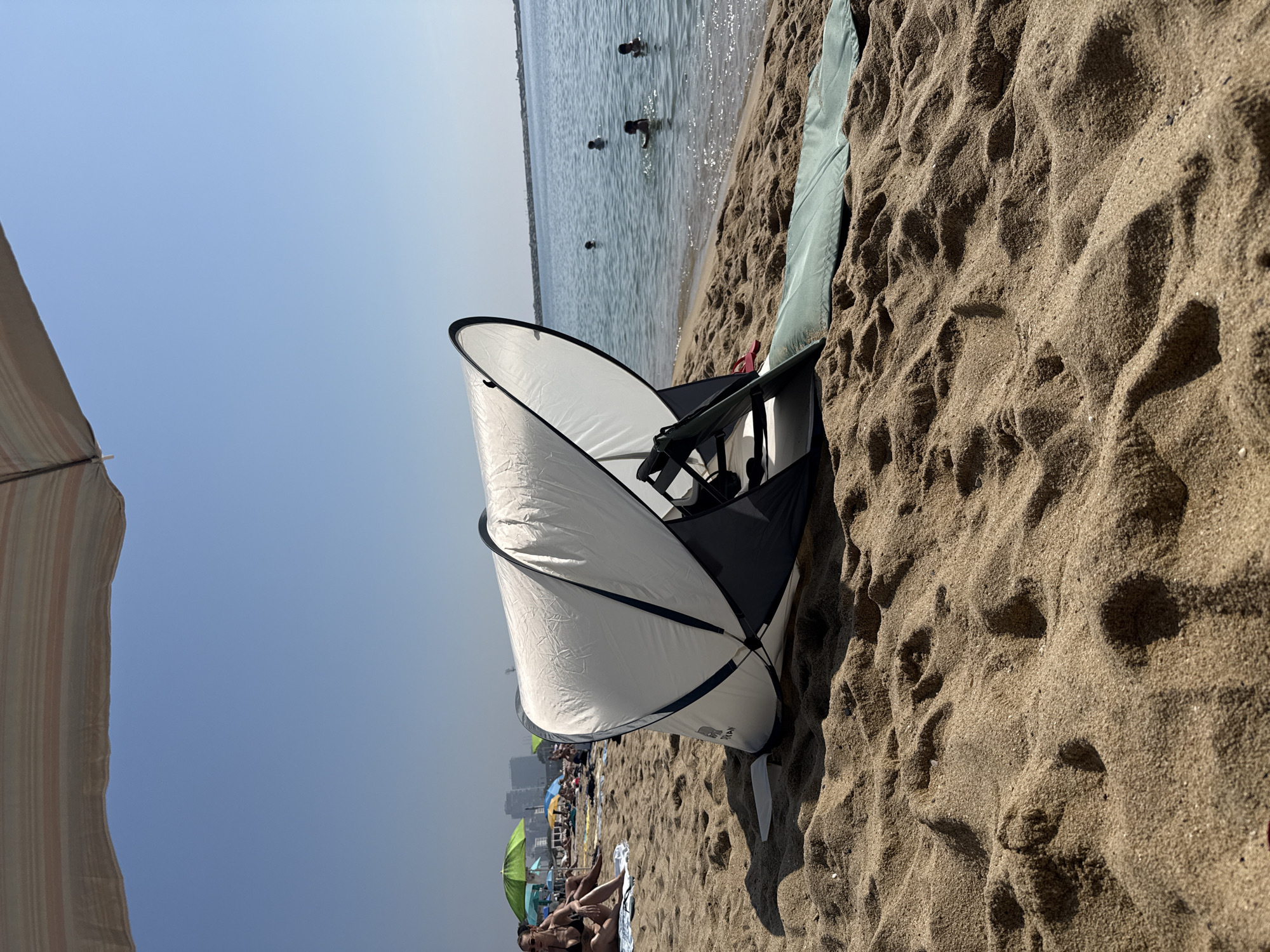 The image depicts a sunny day at a crowded beach in Barcelona, Spain. The foreground is dominated by sand, textured with footprints and shadows, and features a white and dark-colored beach tent. To the left, a portion of a striped beach umbrella is visible against a clear blue sky.
In the middle ground, the gentle waves of the sea meet the shore. Several people are visible in the water, some appearing to be swimming or wading. Further down the beach, numerous beach umbrellas of various colors, including green, are scattered amongst sunbathers and beachgoers. Some people are relaxing on towels, while others are seated or walking along the shoreline. In the distance, a hazy urban skyline suggests the city of Barcelona.
The overall impression is one of a typical summer day at a popular beach, with people enjoying the sun and sea. The clear blue sky indicates fair weather. The orientation of the image is sideways, with the sky appearing on the left and the beach on the right. There is no visible text in the image.