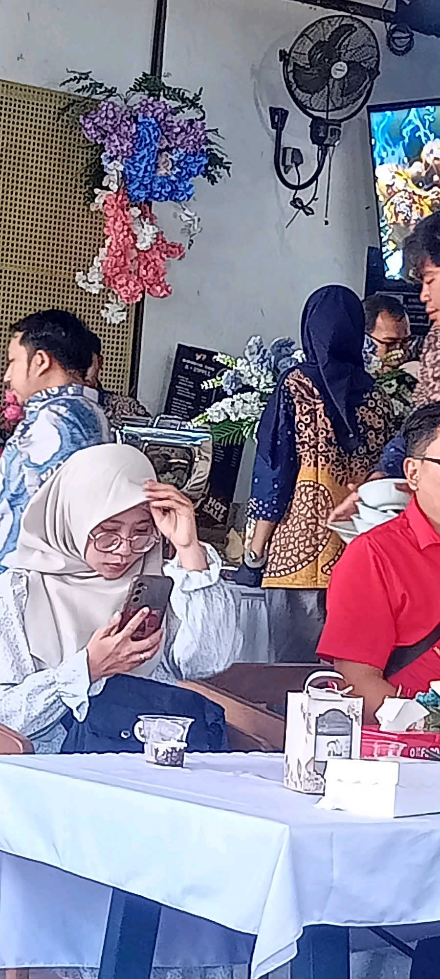 In Desa Ponokawan, Indonesia, a group of people are seated at tables, some engaged with their phones. One woman in the foreground, wearing a light-colored hijab and glasses, is holding a smartphone. Behind her, a man in a patterned shirt is visible. Further back, a person in a dark blue hijab and a patterned outfit stands near a display that reads "HOT SALE". The environment appears to be an indoor seating area, possibly a cafe or restaurant, with decorative floral arrangements and a ceiling fan mounted on the wall. Tables are covered with white tablecloths, and drinks and containers are present on them.