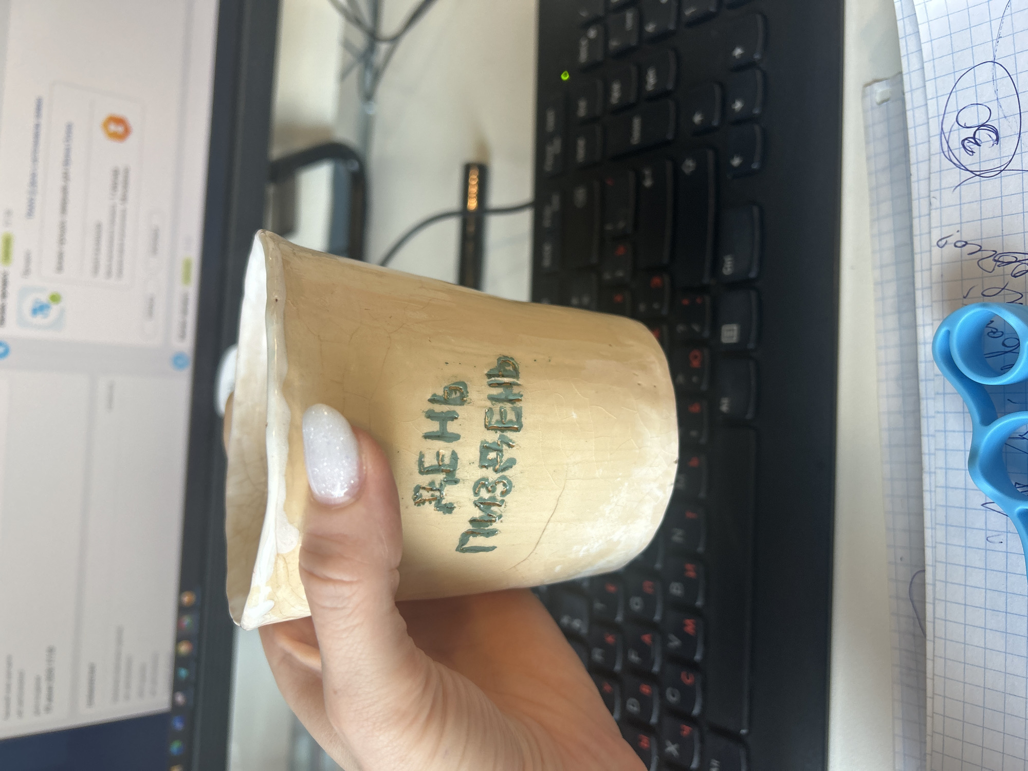 The image shows a person's hand holding a small, light-colored ceramic cup. The cup has a rough texture and appears to be handmade, with visible cracks in the glaze. Dark green lettering is inscribed on the side of the cup. The text on the cup reads "ДЕНЬ ПМЗ, ЕНД". The background suggests an indoor setting, possibly an office, with a computer keyboard visible to the right and a computer monitor to the left. The monitor displays what appears to be a software interface with various icons and text, though the details are out of focus. To the far right, a sheet of graph paper with a hand-drawn circle containing the number "30" in blue ink is visible, along with a pair of blue scissors. The lighting appears to be artificial, consistent with an indoor environment. No specific time of day or weather conditions can be inferred. The overall impression is of a casual moment captured at a desk.