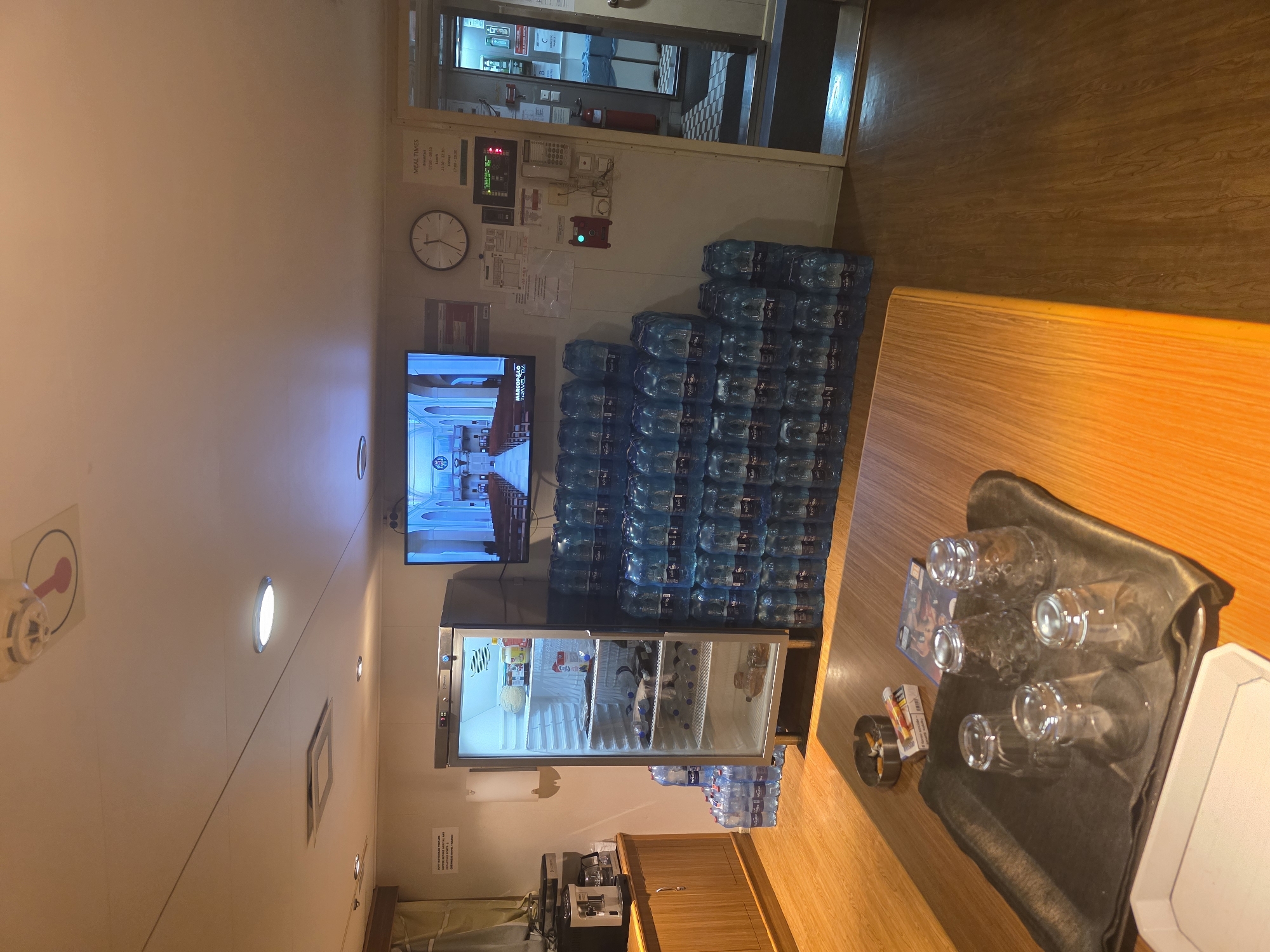 The image depicts the interior of a common area, possibly on a ship or in a facility, given the arrangement of supplies and equipment. The scene is well-lit by overhead lighting and the illuminated television screen.
Dominating the right side of the room is a large stack of blue plastic water bottles, neatly arranged in a pyramid formation. To the left of the water bottles, a tall, stainless-steel refrigerator is open, showcasing its contents, which include various food items and beverages. Next to the refrigerator, a dark wooden cabinet sits below a coffee machine.
A wooden table is positioned in the foreground, on which rests a black tray holding several clear glasses. An ashtray with cigarette butts and a pack of cigarettes are also on the table, along with a video game case.
On the wall, a flat-screen television displays an image of church pews and stained-glass windows, suggesting a religious broadcast. Above the television, a clock is mounted on the wall. A sign with "MEAL TIMES" is also visible. Further down the wall, a red fire alarm and an intercom system are mounted. An open doorway on the right side of the image leads to another area, partially obscured, with a checkered floor pattern.
The environment appears to be a utilitarian space, perhaps a break room or a mess hall. The presence of a large quantity of water bottles suggests a need for hydration, possibly in a location where fresh water is scarce or for a group of people. The overall impression is one of practicality and provision. There are no people visible in the image.
The location is not the city of Brindisi, Italy.