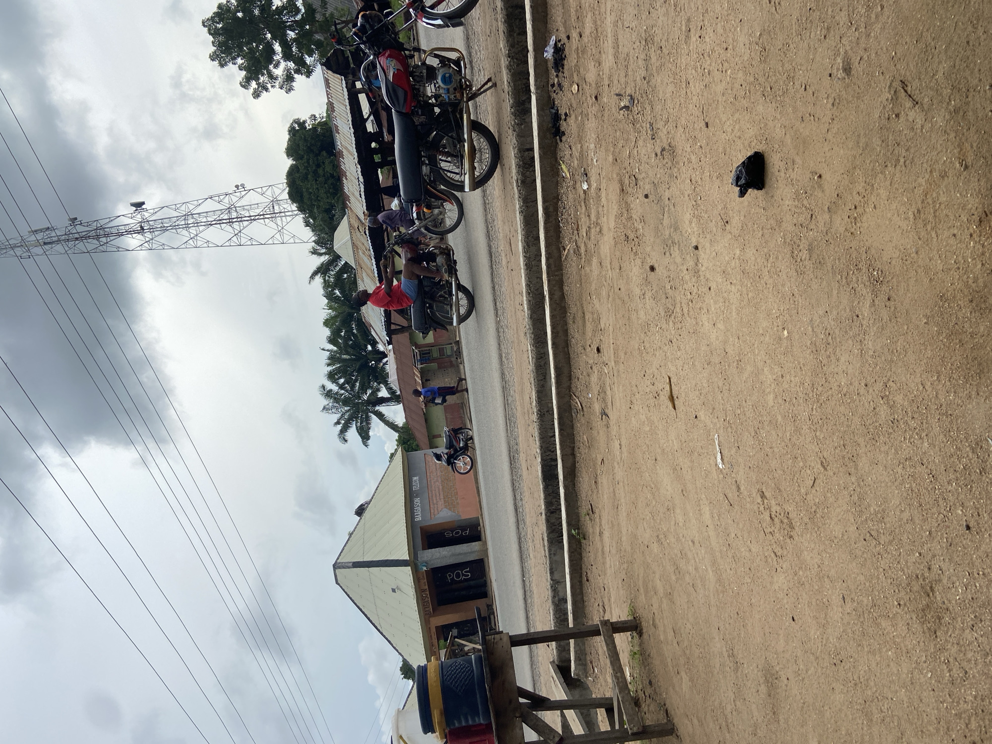 The image shows a street scene in Mararaba, Nigeria. The sky is overcast. There are multiple motorcycles parked along the side of the road. A man wearing a red shirt is on one of the motorcycles. A building labeled "BARAGASON TELECOM" is visible, with "POS" signs displayed. An electrical transmission tower stands in the background. A dirt area is visible on the right side of the image, with a piece of black debris. Electrical wires run across the sky. Palm trees are visible. A wooden structure is visible in the lower right corner.