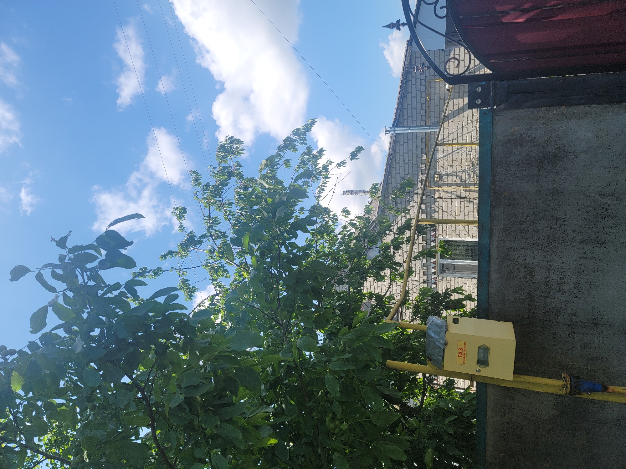 In the city of Vovkivka, Ukraine, a bright yellow gas meter box is mounted on a yellow pipe attached to a grey concrete wall. The box has a metal lid covered with a grey, crinkled material. Text in Russian, reading "ГАЗ" and "ВОЗГНЕБЕЗОПАСНО" (meaning "GAS" and "FIRE HAZARDOUS"), is visible on the front of the box. The gas meter is positioned under a large green tree with numerous leaves, its branches extending towards the sky. Behind the wall, a light-colored brick building with a white-framed window is visible. A yellow metal railing runs horizontally in front of the building. The sky above is a clear blue, dotted with white, fluffy clouds, and several power lines are strung across it. To the right, a portion of a dark red metal gate with decorative ironwork is visible.