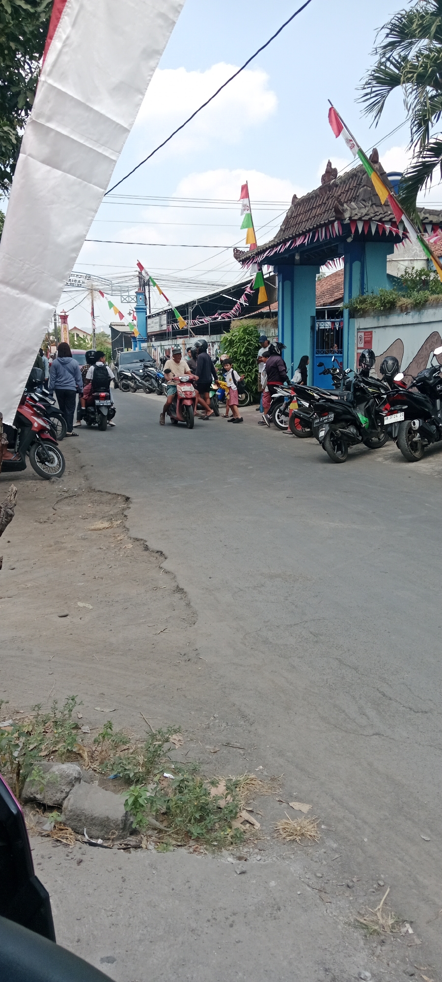 The image depicts a street scene in Purwomartani, Indonesia, likely on a day of celebration or a public event. A prominent white banner hangs from the left side, partially obscuring the view. The street is paved with asphalt, showing signs of wear and tear with a large pothole on the left side of the foreground.
A number of people are gathered on the street, primarily interacting around motorcycles. Several motorcycles are parked or being ridden. The architecture in the background suggests a local community, with a blue building featuring a traditional Indonesian roof design. Decorative flags, predominantly red and white (the colors of the Indonesian flag), are strung across the street and adorning the entrance to the blue building, indicating a festive atmosphere, possibly related to Indonesian Independence Day.
On the left side of the image, several people are standing or walking near parked motorcycles. In the center, a man is riding a pink scooter, and other individuals are standing around him. To the right, more people and motorcycles are visible, with some individuals walking towards the blue building. A child in a red shirt and shorts is walking with an adult near the entrance. The environment includes trees and vegetation along the side of the road. The sky is cloudy.