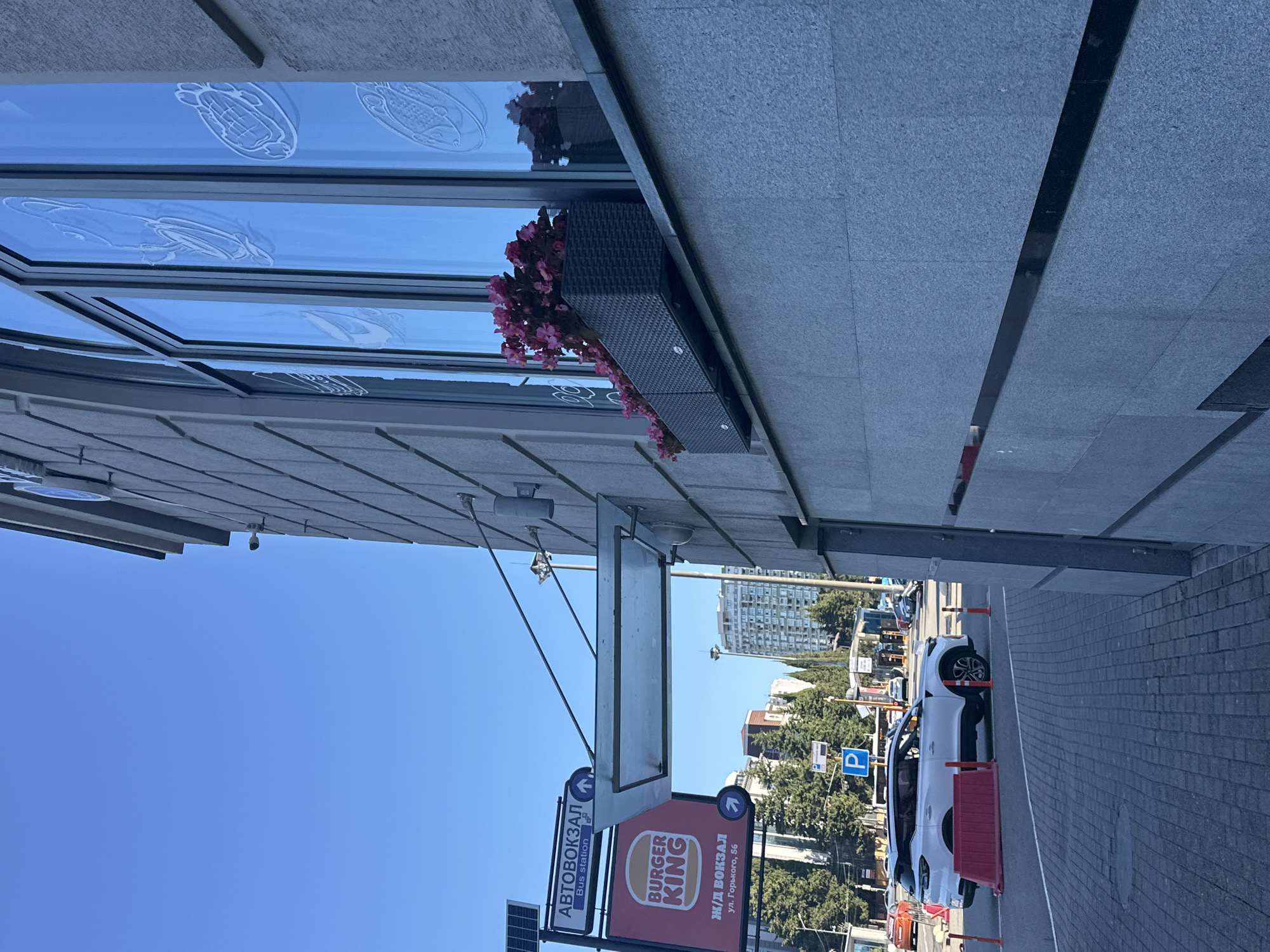 The image is taken from a low angle, looking up at the facade of a modern building in Sochi, Russia. The building has large, reflective windows with stylized etched images of food items. A planter box filled with pink flowers sits on the windowsill. To the left, a bus station sign is visible, along with a Burger King sign. A white SUV is parked on the street, with orange traffic cones and a red barrier in the foreground. The sky is clear and blue. The overall scene suggests a public urban space with commercial establishments and transportation infrastructure.