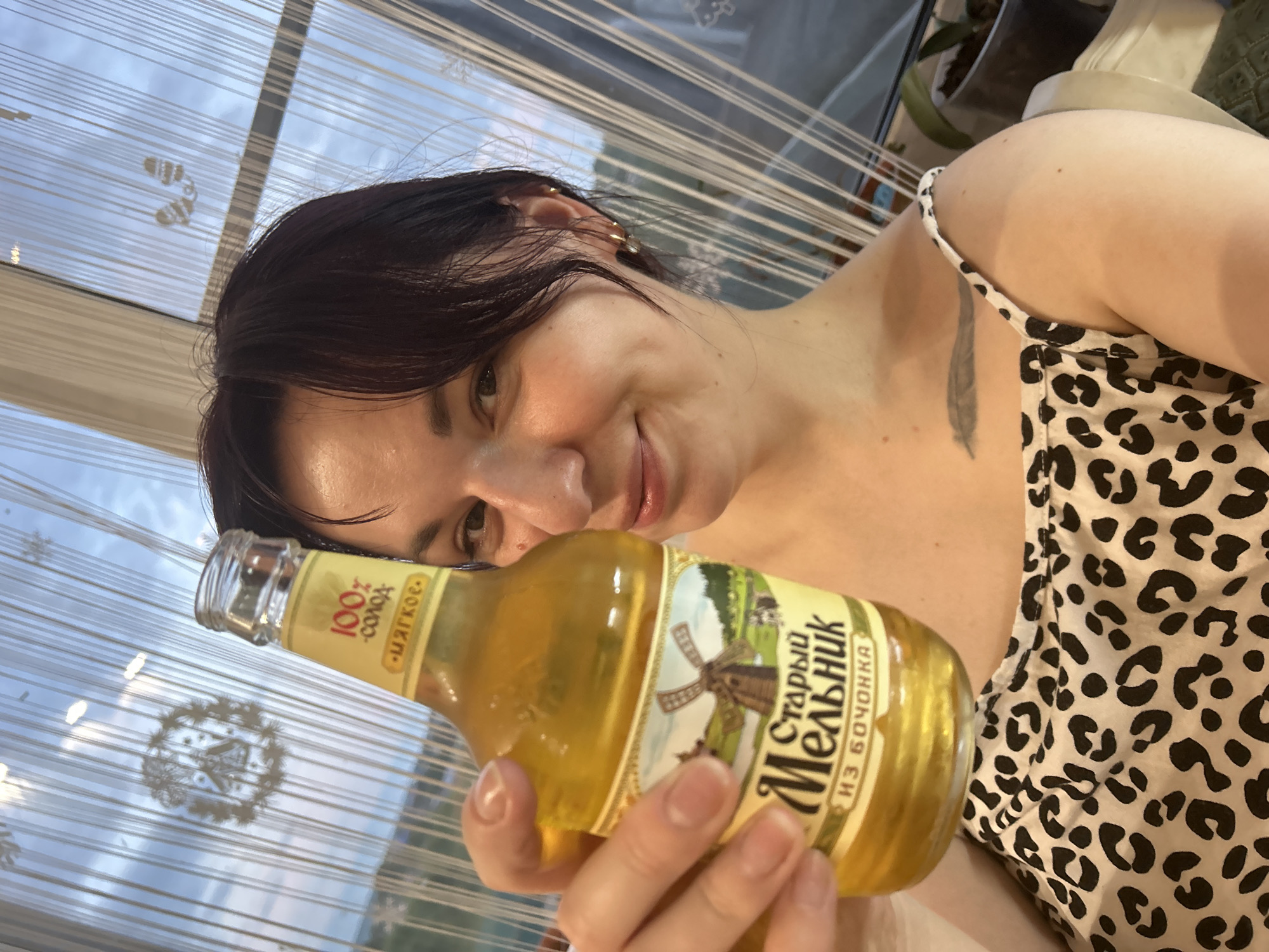 A young woman with dark hair is taking a selfie while holding a bottle of beer in her right hand. She is wearing a leopard-print top. The background consists of a window with vertical blinds and the sky outside. The beer bottle has a yellow label with text and an image of a windmill. The scene appears to be indoors, possibly in a residential setting in the city of Fokino, Russia.