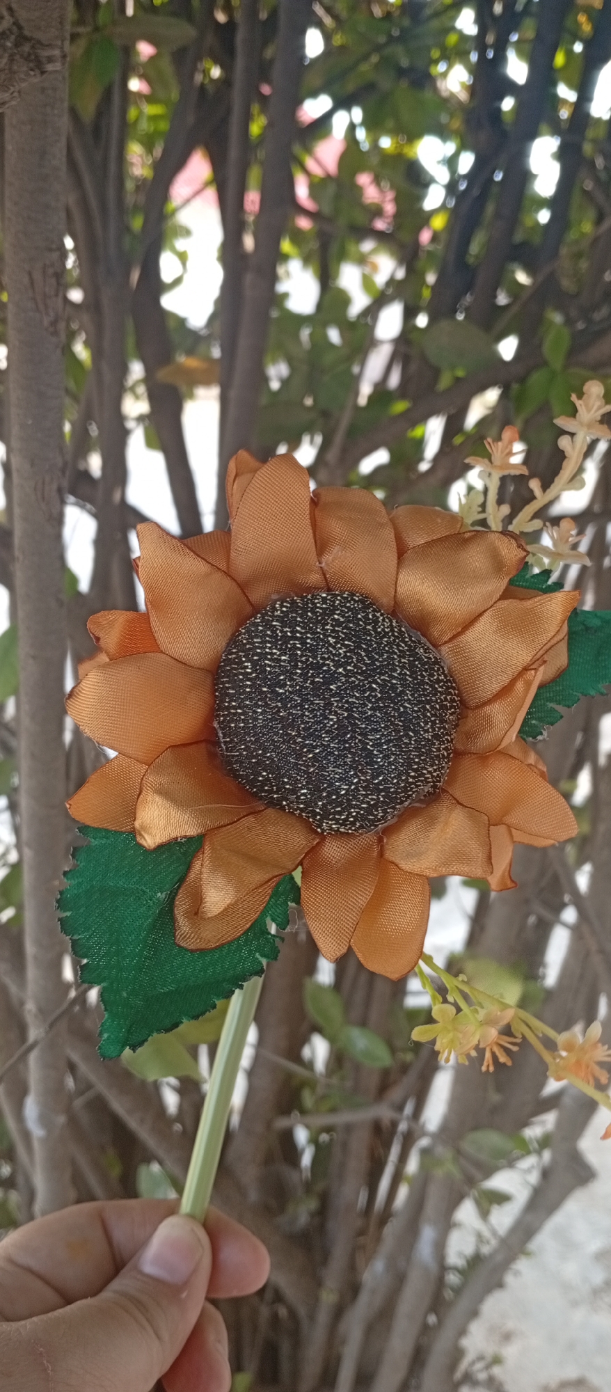 The image presents a close-up, vertical shot of an artificial sunflower held by a person's hand against a blurred outdoor background, likely taken in Juan Griego, Venezuela.
The primary subject is a vibrantly colored artificial sunflower. Its petals are made of shiny, orange fabric, arranged in several layers. The center of the sunflower is dark brown or black, with a textured, speckled pattern that mimics the seeds of a real sunflower. A single green fabric leaf, with jagged edges, is attached to the flower's light green stem. To the right of the sunflower, a small cluster of pale yellow or light orange artificial flowers with tiny, delicate stamens is also visible.
A human hand, with a light skin tone, holds the stem of the artificial flower near the bottom of the frame, with fingers gently curved around it. The background is composed of out-of-focus trees and bushes, featuring slender dark trunks and branches, interspersed with glimpses of green foliage and bright spots of sunlight filtering through, indicating it is daytime. The soft, blurred background helps to draw attention to the sharply focused artificial flower. There is no visible text in the image. The scene suggests a bright, pleasant outdoor setting.