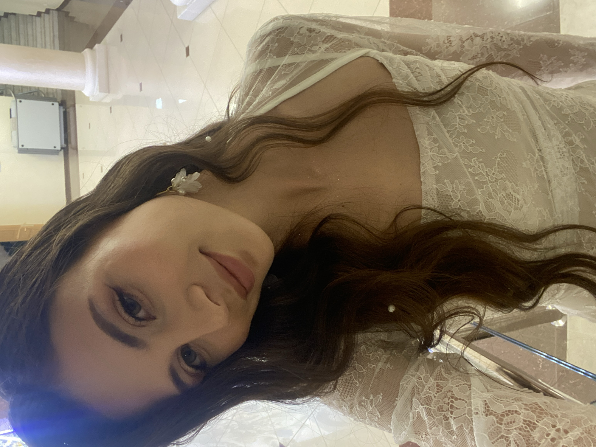 The image features a close-up, sideways selfie of a woman. She has long, dark, wavy hair with some strands falling over her face. She is wearing a white lace garment, possibly a dress or robe, with intricate floral patterns. There are small pearl-like embellishments in her hair and on her ear. Her makeup includes eyeliner and mascara, with a soft pink lipstick. She is looking towards the camera with a gentle expression.
The background is blurred but suggests an indoor setting with light-colored walls and flooring. There are architectural elements visible, such as a column and what appears to be part of a door or cabinet on the left. The lighting is bright, indicating it could be daytime or well-lit indoors. There is no visible text in the image. The context provided states the location is Votkinsk, Russia, but there are no specific visual cues in the image to confirm this. The overall mood is soft and intimate.