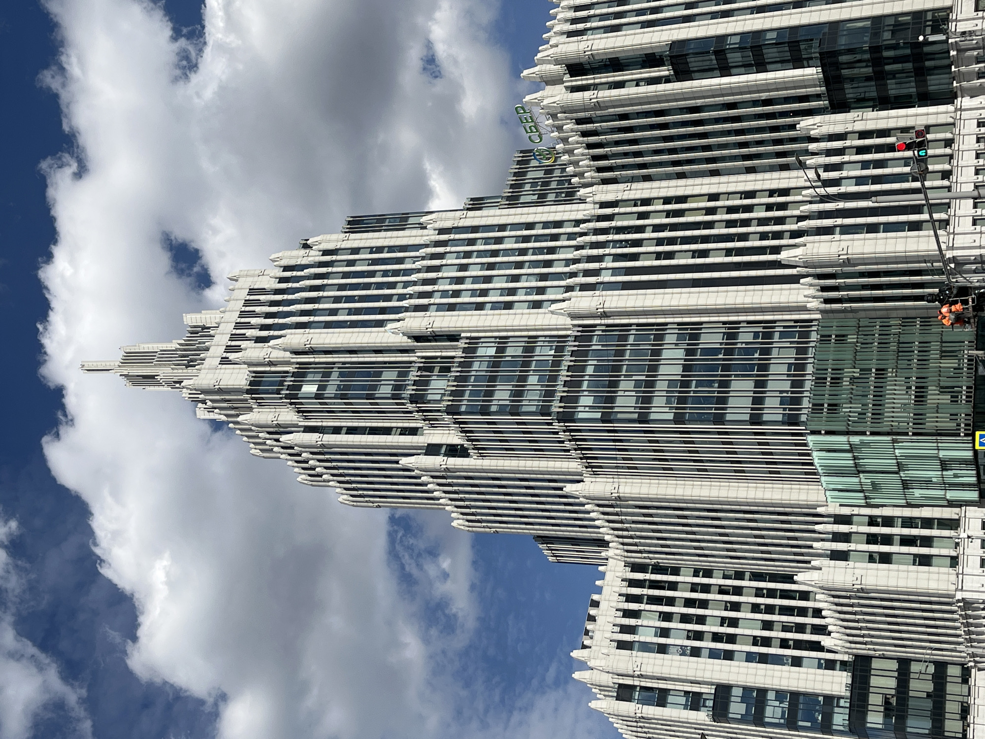 The image shows a modern skyscraper in Moscow, Russia, viewed from a low angle. The building has a stepped, ziggurat-like design, with many horizontal lines formed by its architecture. A green "Cбер" (Sber) sign is visible on the upper portion of the building. A traffic light pole with signals is present near the building's facade. The sky has a mix of blue and white clouds.