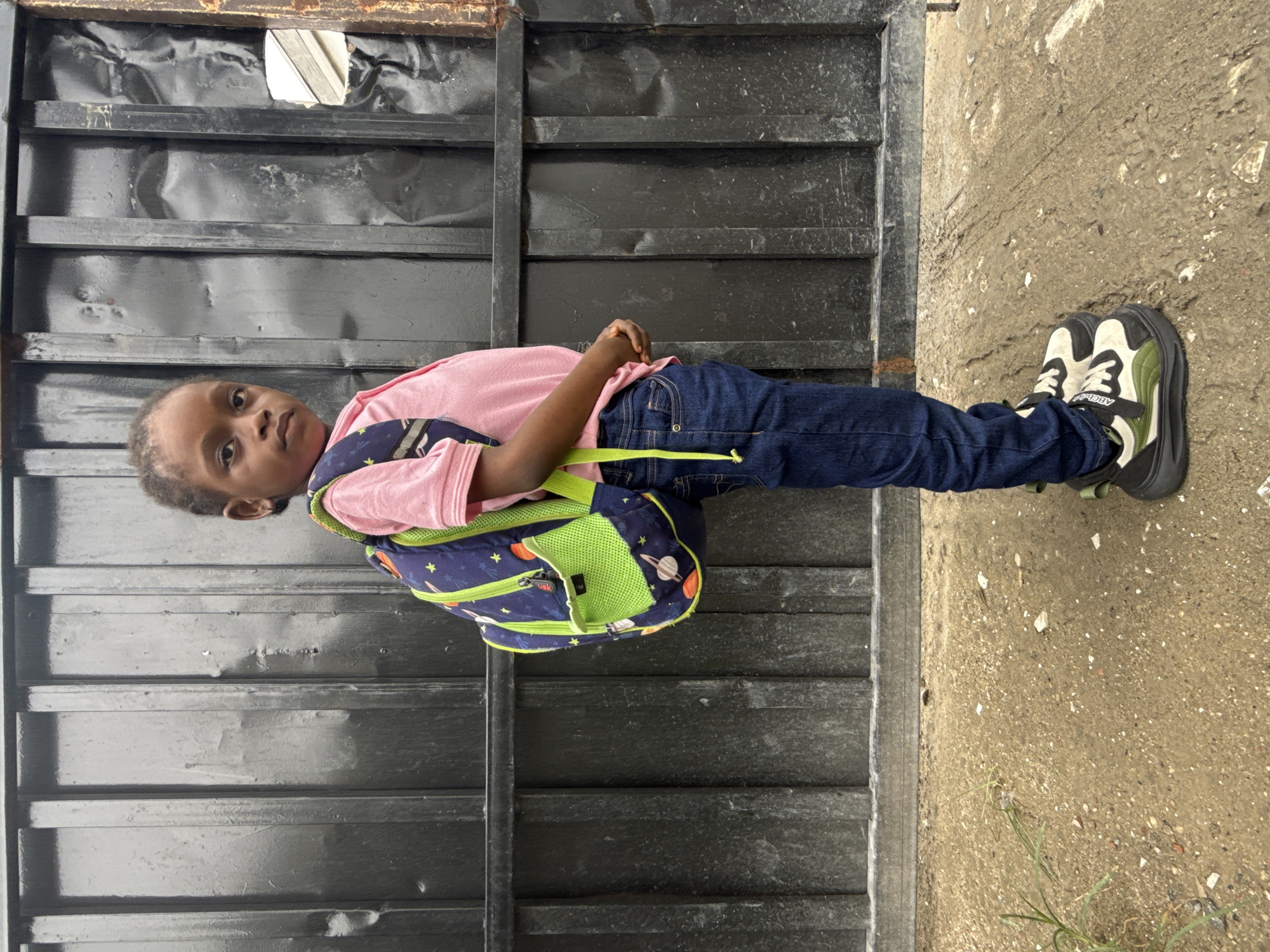 A young child stands facing left in front of a black, metal corrugated door. The child wears a light pink shirt, blue jeans, and black, white, and green sneakers. A space-themed backpack is slung over the child's shoulders. The child looks toward the viewer. To the right of the metal door is a concrete surface with debris on it. The location is Ado, Nigeria.