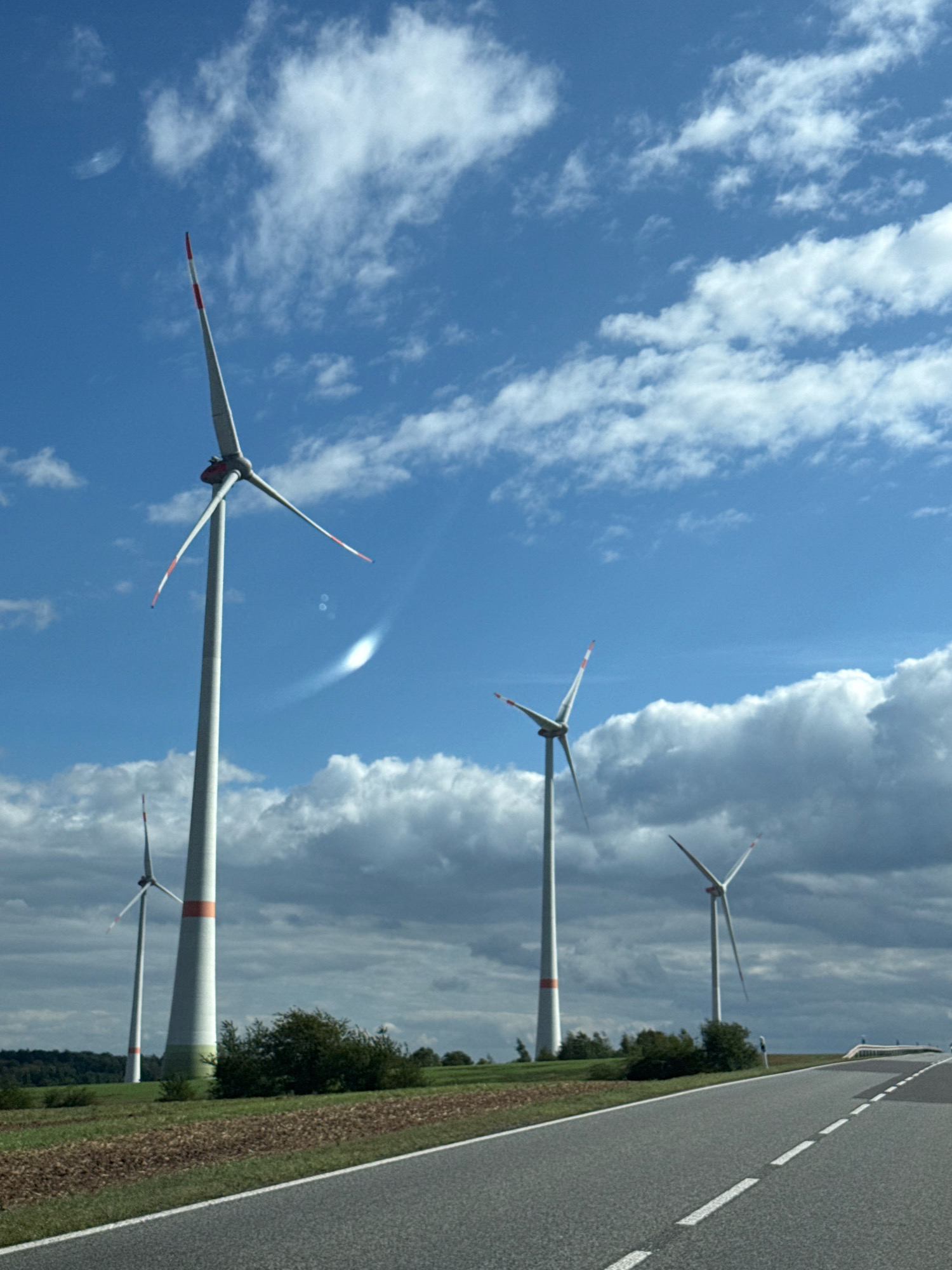 The image depicts a landscape scene featuring several wind turbines against a bright blue sky with scattered white clouds. A paved road with white dashed lines runs diagonally from the lower right to the lower left of the frame. The ground alongside the road is a mix of tilled soil and green grass. In the background, more wind turbines are visible, along with a treeline and some low-lying bushes. The overall impression is of an open, rural environment, likely a wind farm in Germany, given the context of Kleinfurra. There are no people or animals visible in the image.